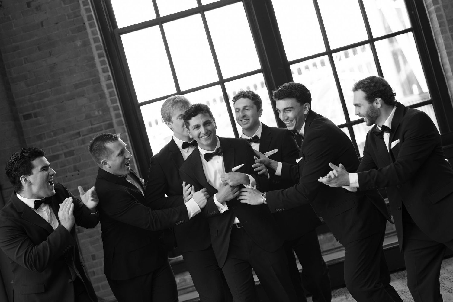 Groomsmen in black tuxedos laughing and celebrating together in a black and white wedding photo.