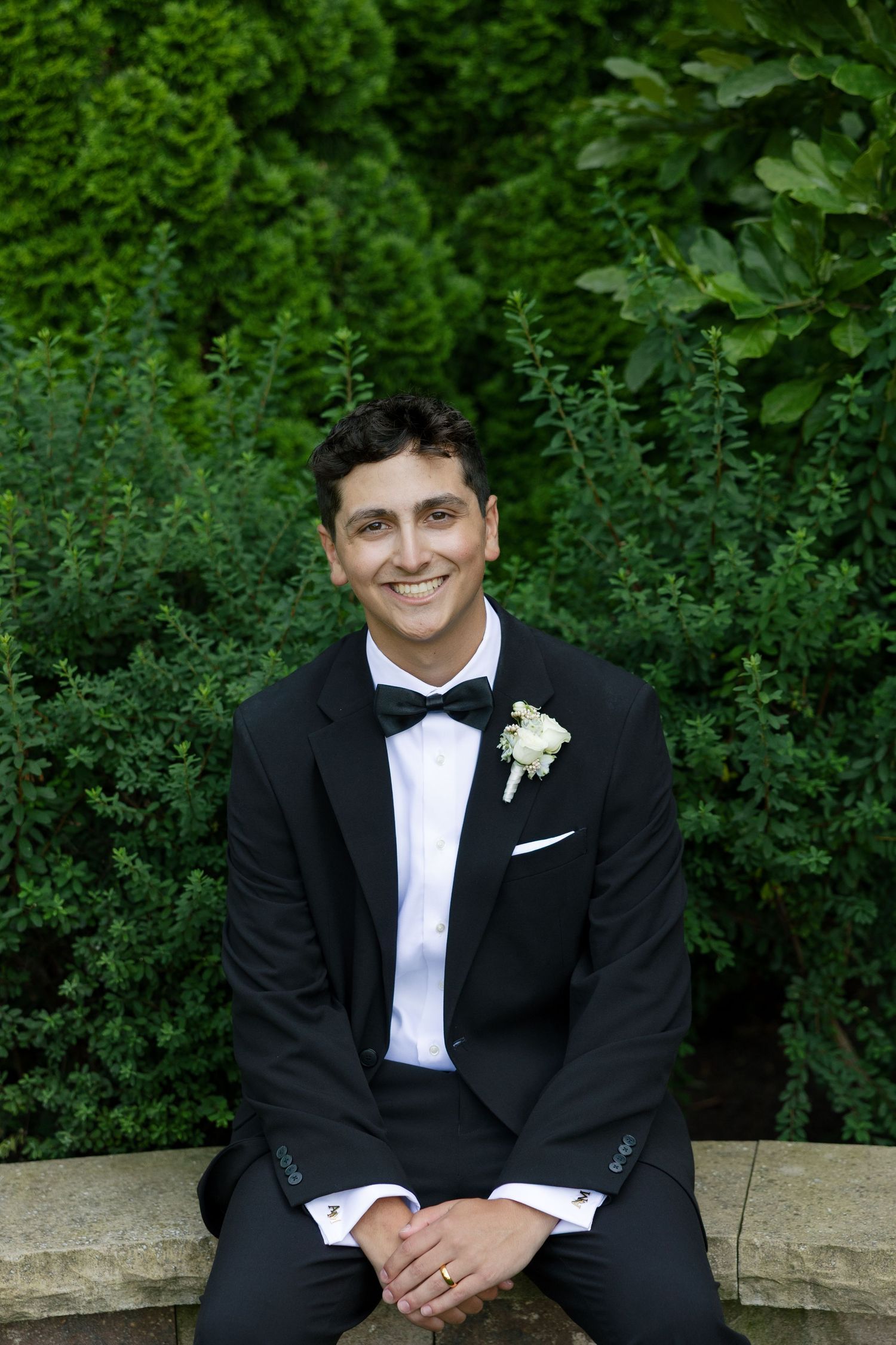 Handsome groom in black tuxedo with bow tie and boutonniere sitting outdoors in front of lush green hedges.