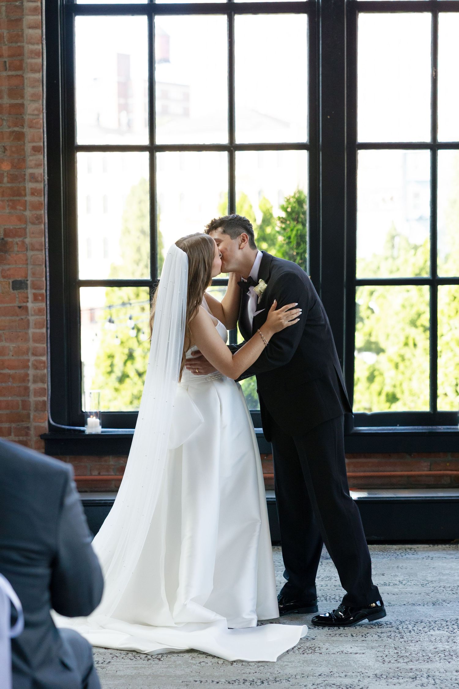 Bride and groom sharing first kiss as newlyweds before large industrial windows in elegant brick ceremony venue.
