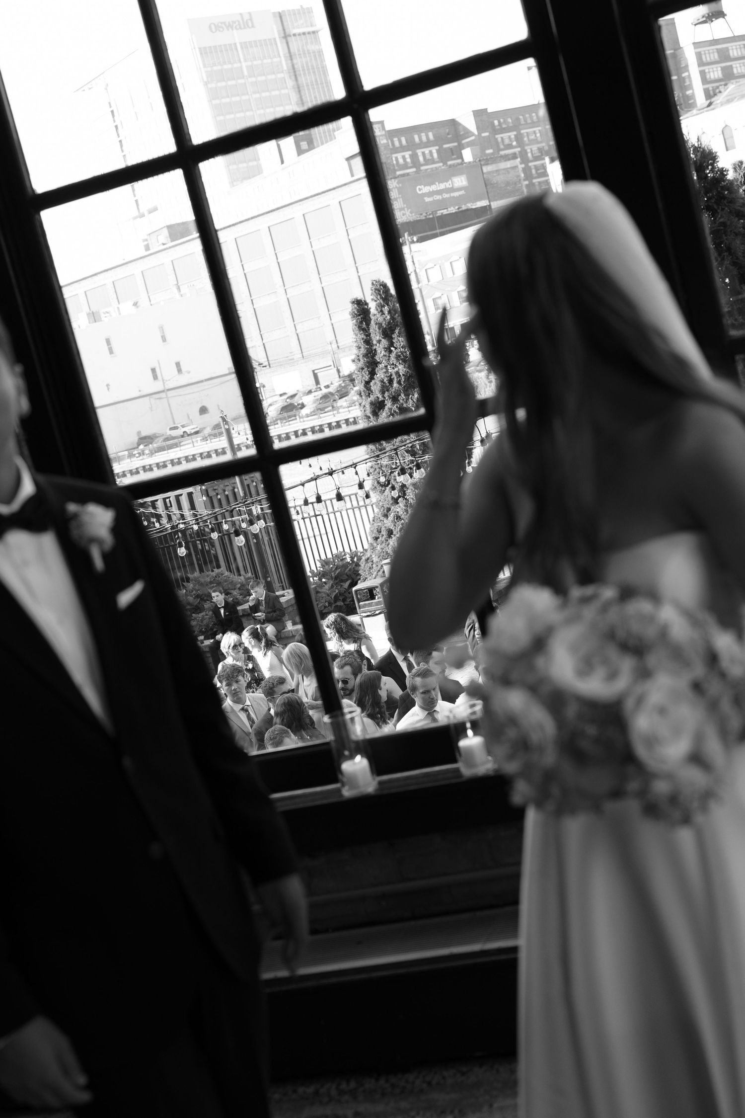 Black and white photo of bride and groom peering through large window at wedding guests gathered outside venue.