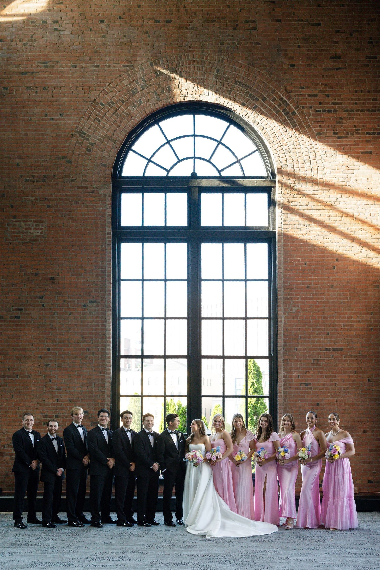Wedding party posing before arched window in brick venue, bridesmaids in pink gowns and groomsmen in black tuxedos.