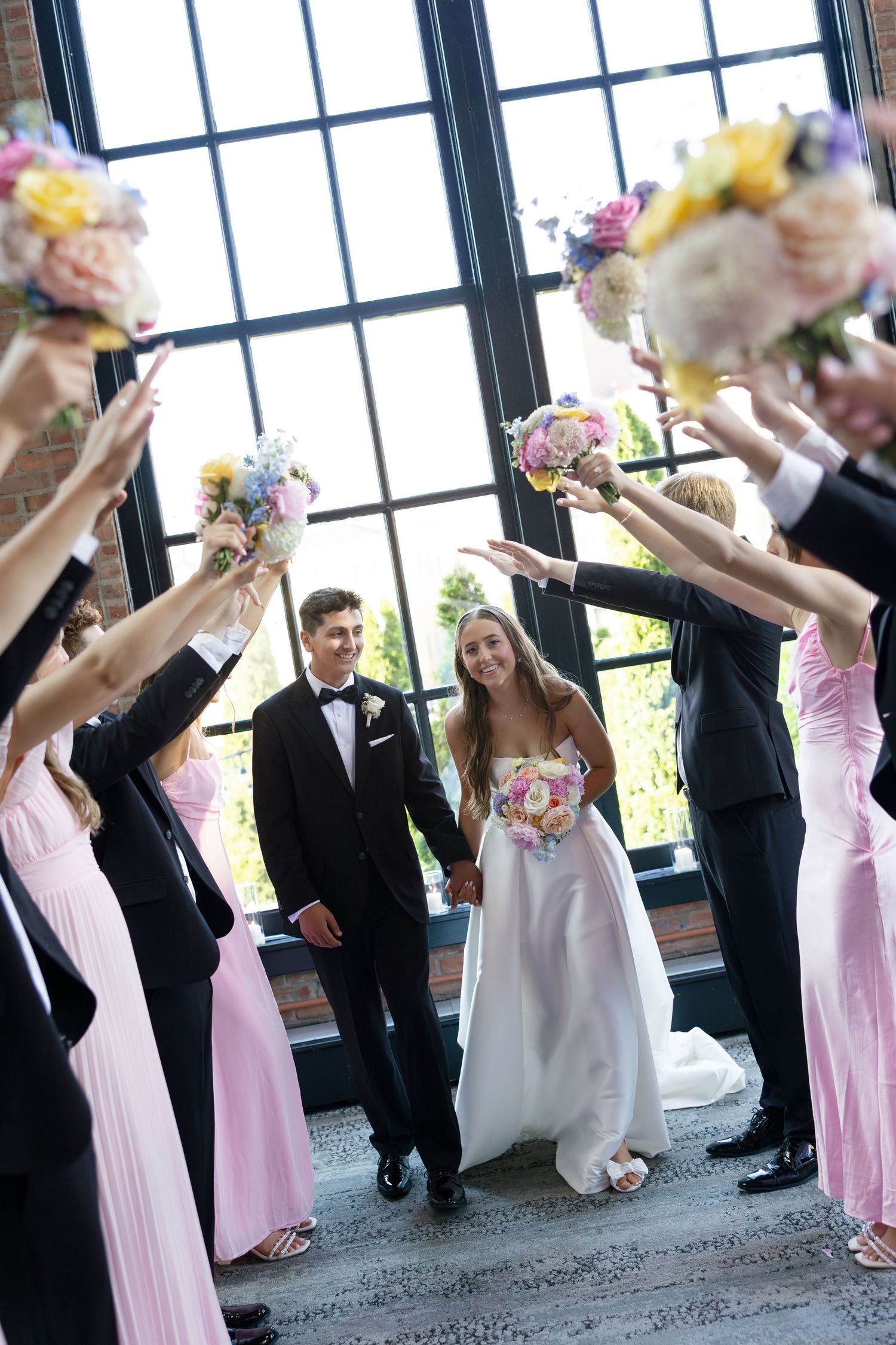 Bridal party raising colorful bouquets to create floral arch as bride and groom walk through in brick venue.