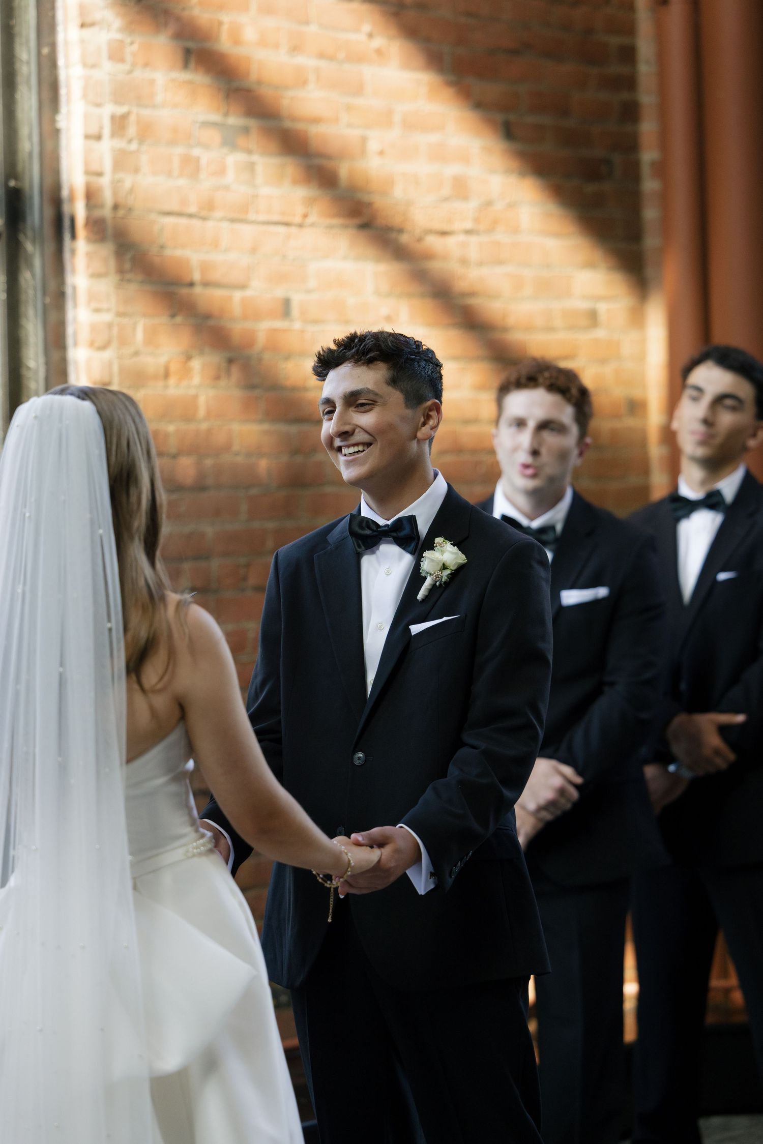 Groom smiling at bride during wedding ceremony in brick venue, with groomsmen standing behind in black tuxedos.