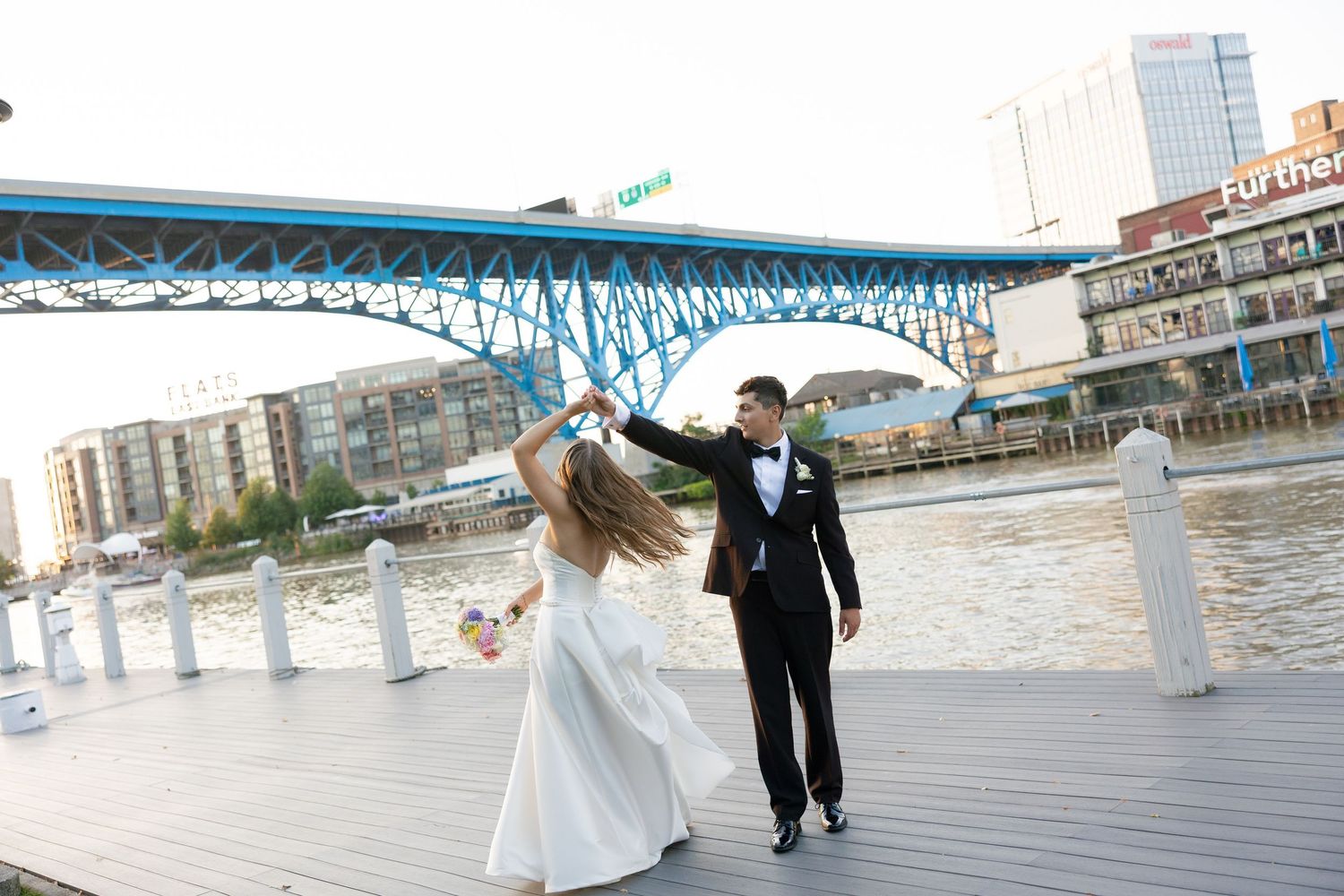 Bride and groom dancing joyfully on waterfront dock beneath iconic blue bridge on sunny wedding day.
