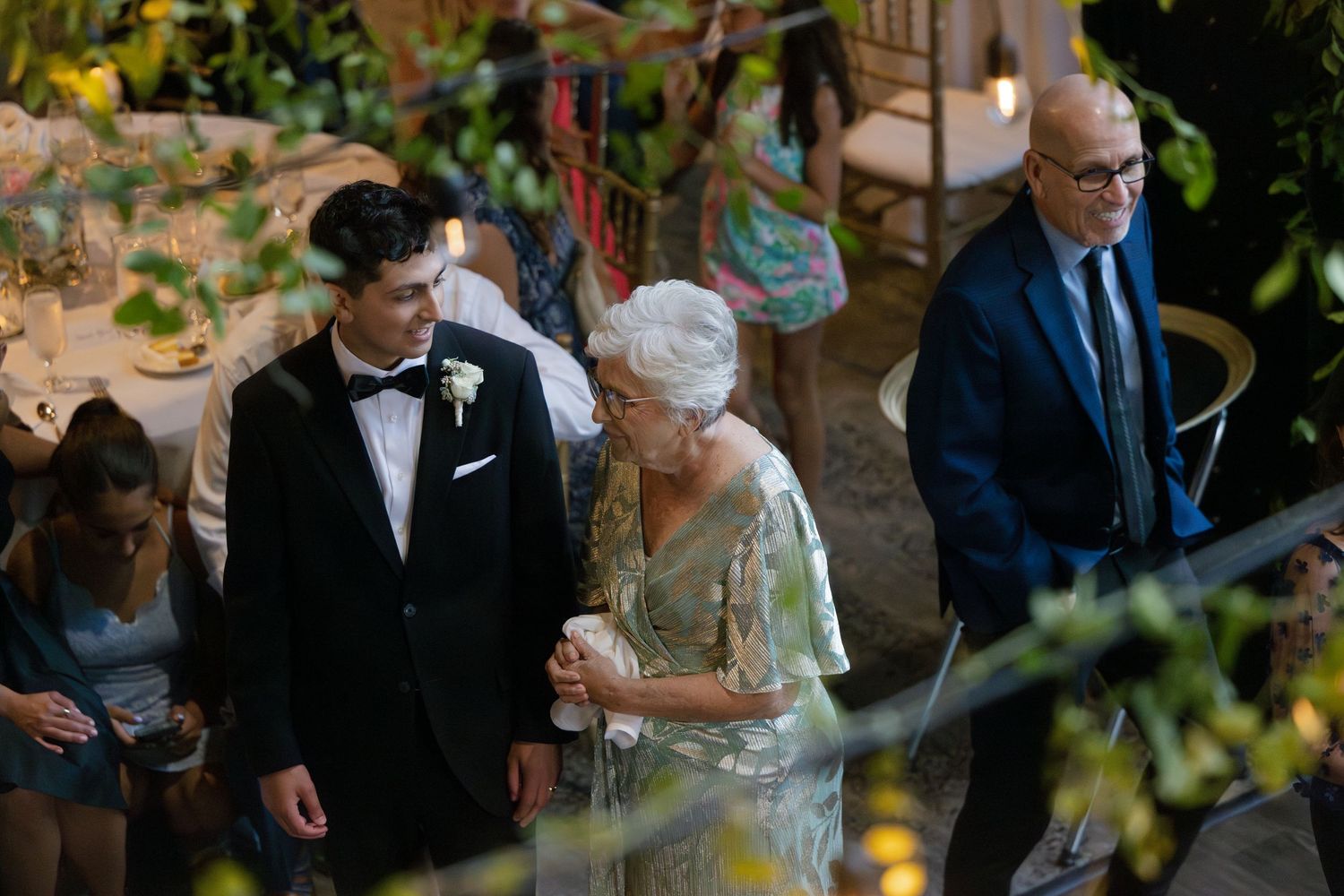 Groom in black tuxedo escorts elderly woman in elegant gown through lush garden wedding venue.