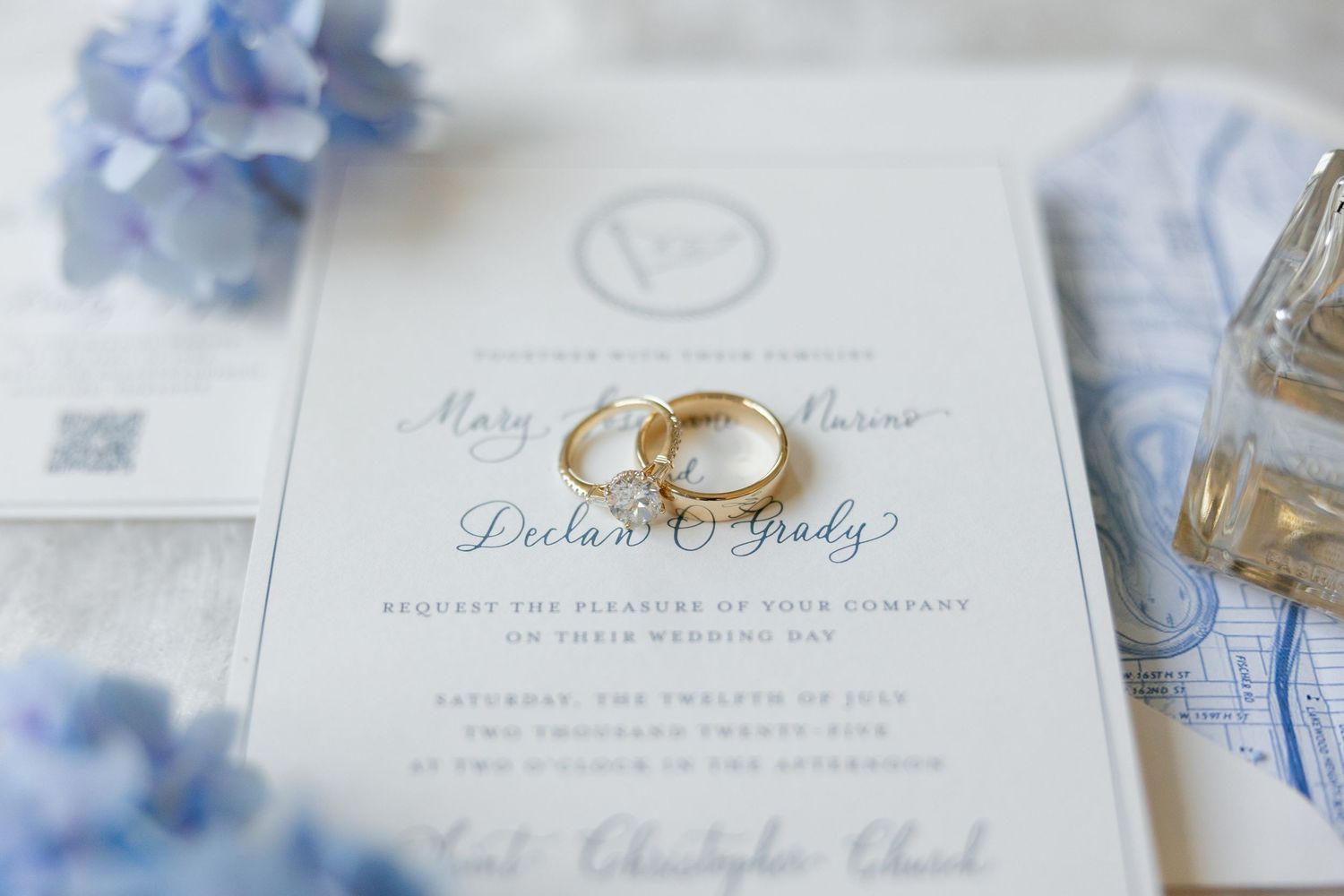 Elegant wedding invitation suite with gold rings, blue hydrangeas, and perfume bottle.