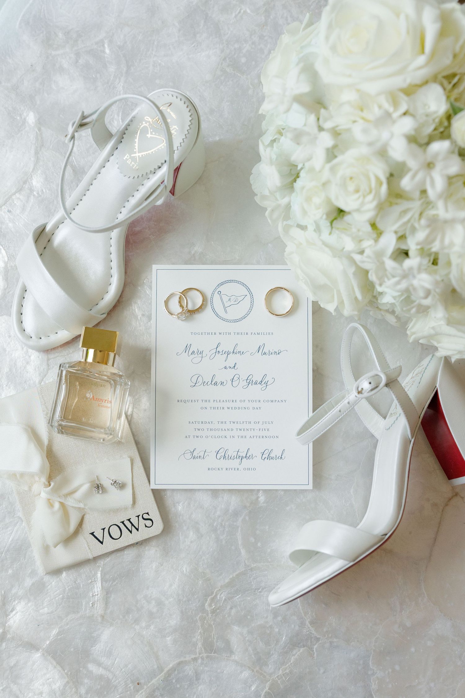 Wedding flat lay with white heels, invitation, gold rings, perfume, and white floral bouquet.