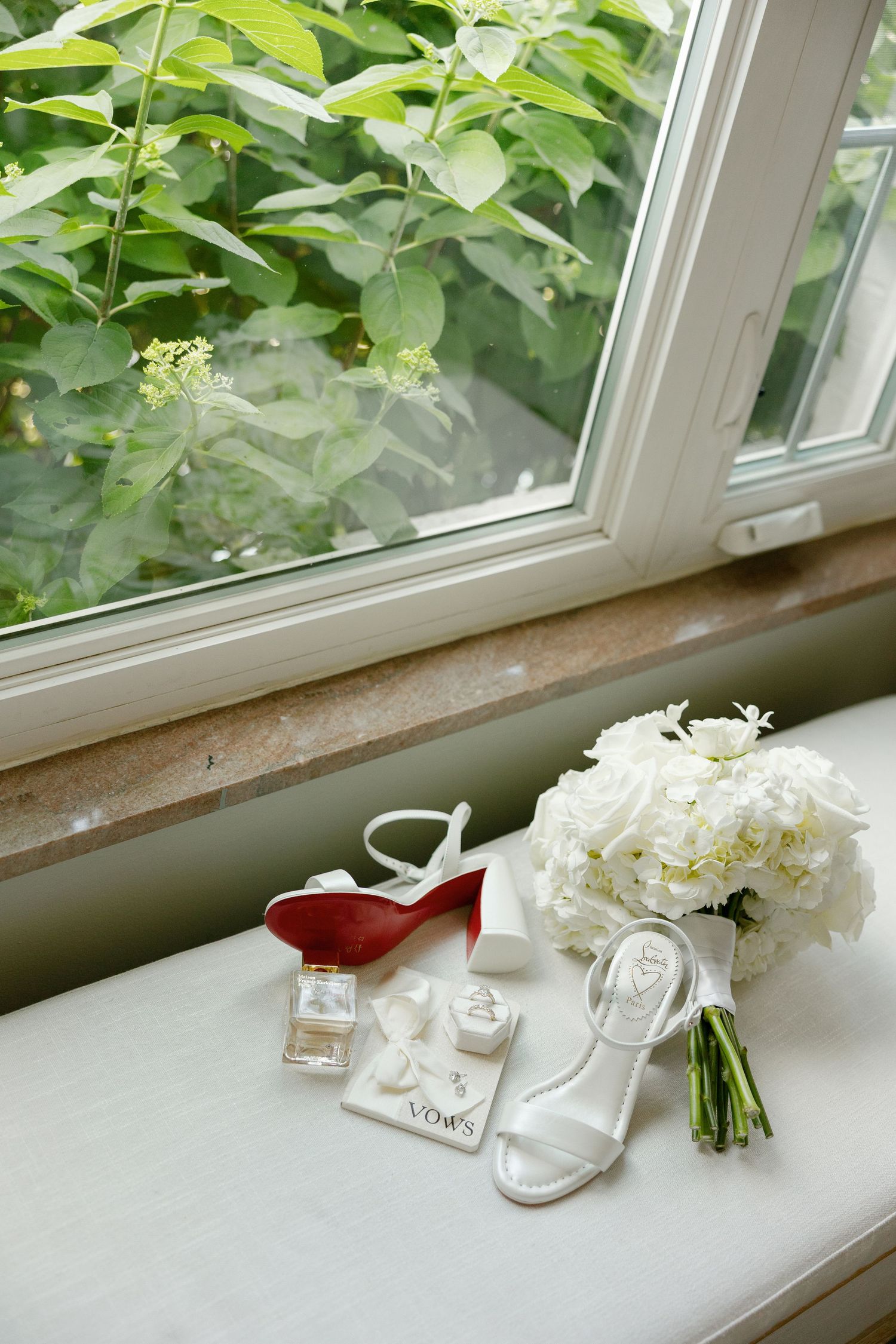 Bridal accessories including white heels, red shoes, bouquet, and perfume on window sill.
