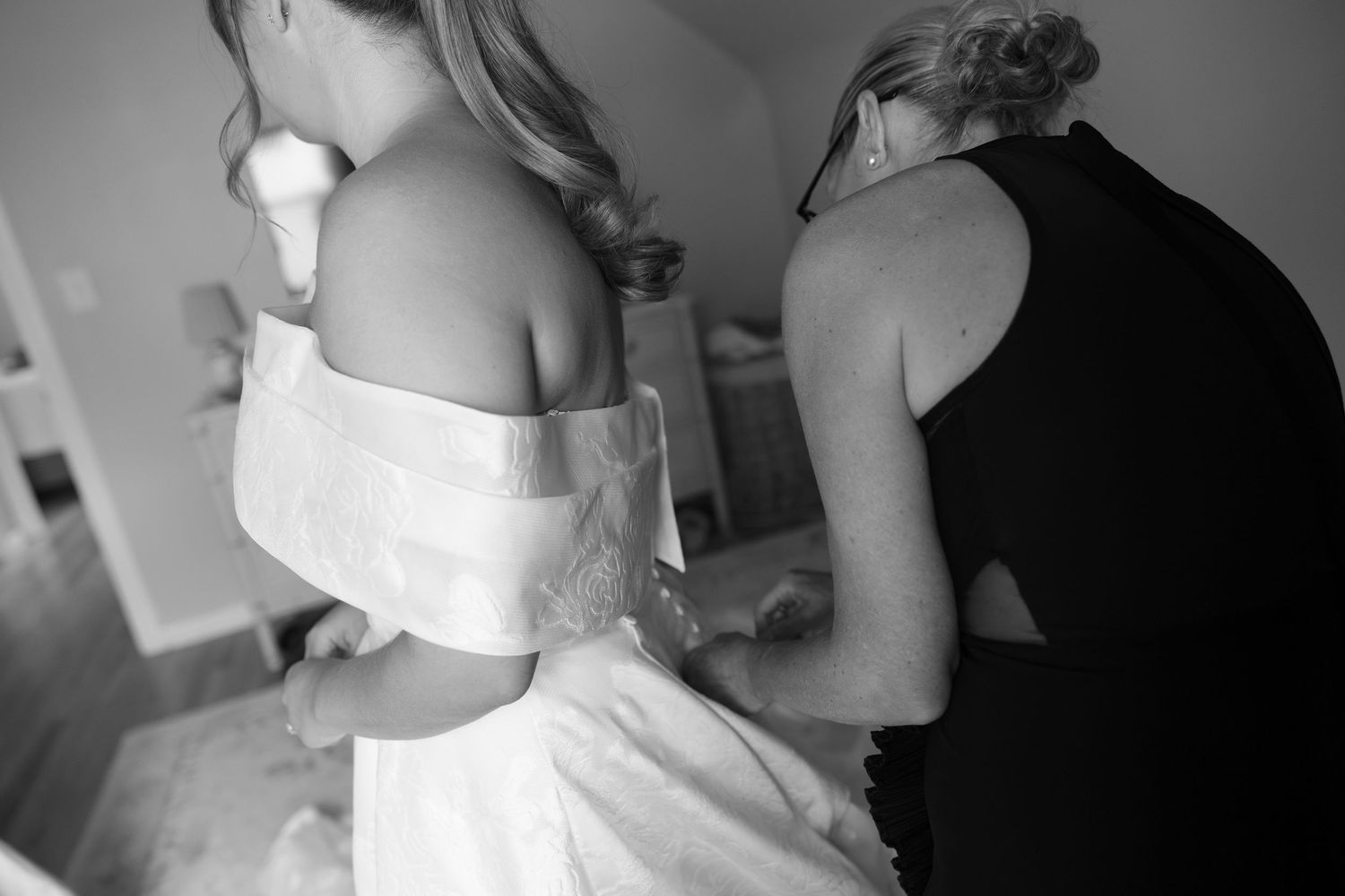 Helper fastening the back of a brides white wedding gown in black and white photo.