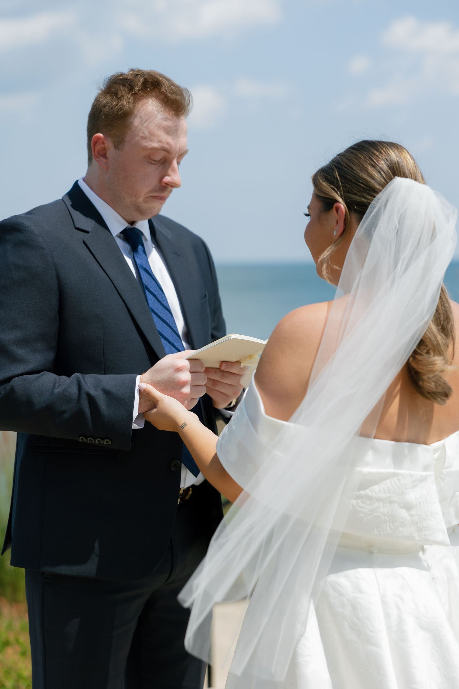Groom reading wedding vows to bride outdoors by the ocean on a sunny day.
