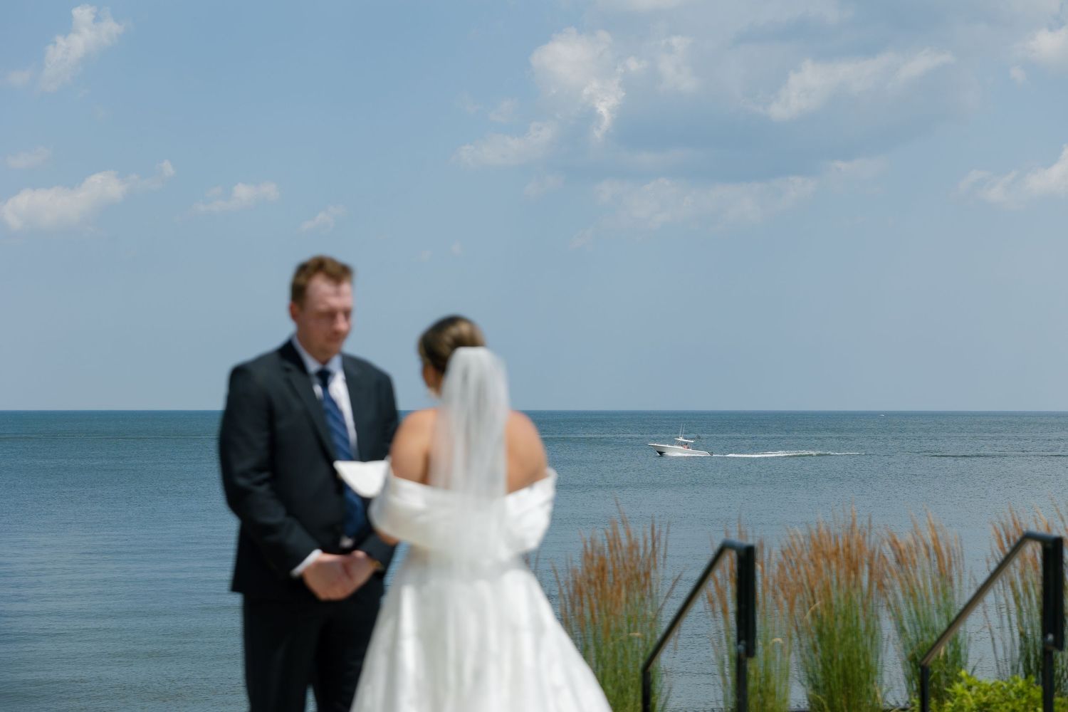 Bride and groom exchanging vows at an outdoor waterfront ceremony with a boat passing by.