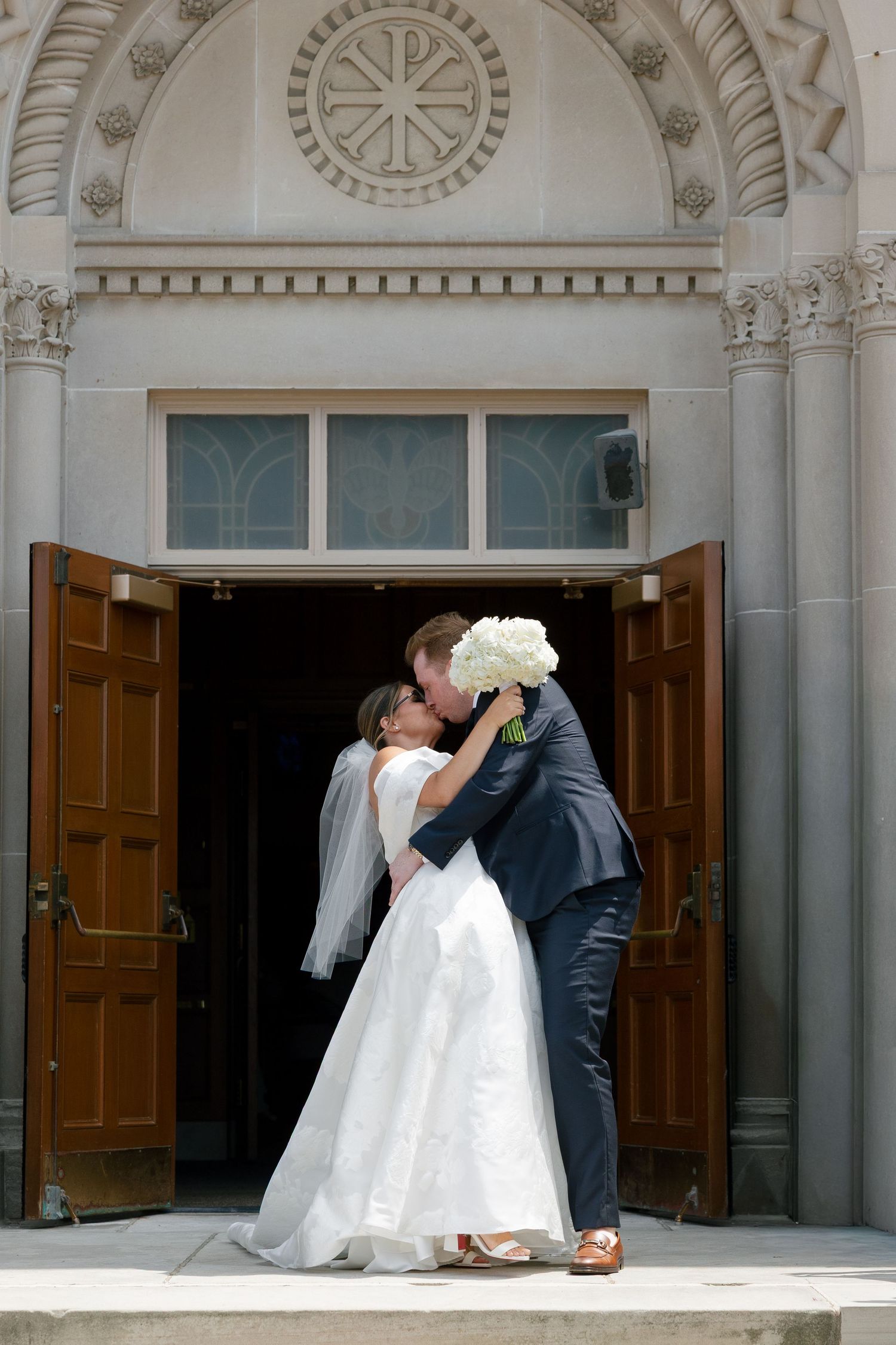 Bride and groom sharing a kiss at the entrance of an ornate stone church.