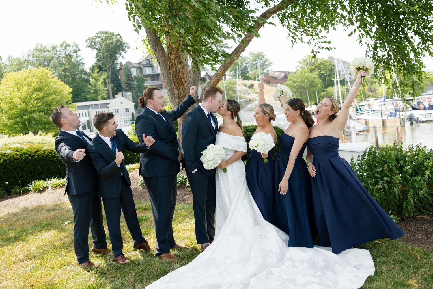 Wedding party celebrating with the kissing couple near a marina under a shady tree.