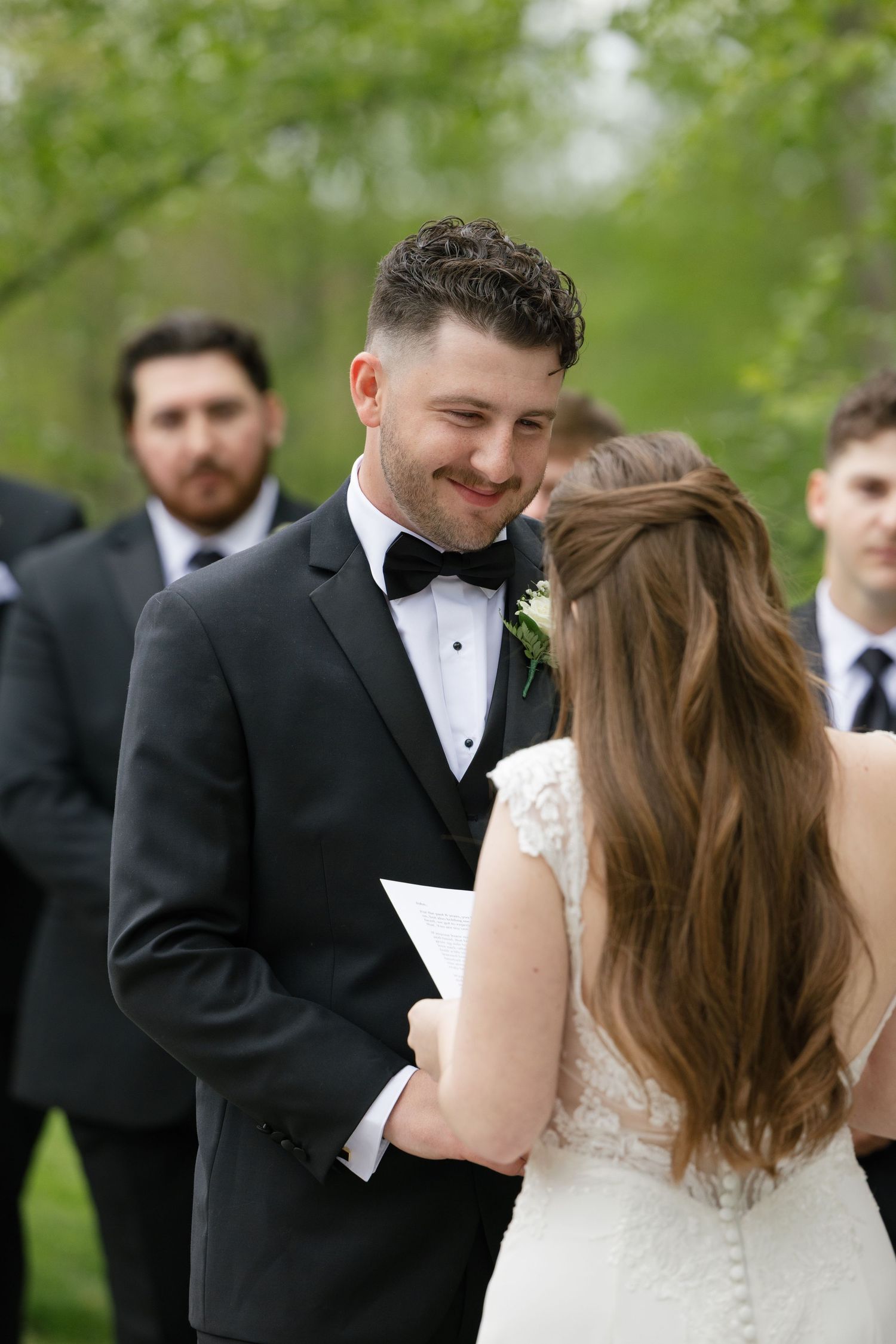 A smiling groom in a black tuxedo reads his vows to his bride during an outdoor wedding ceremony in a green park.