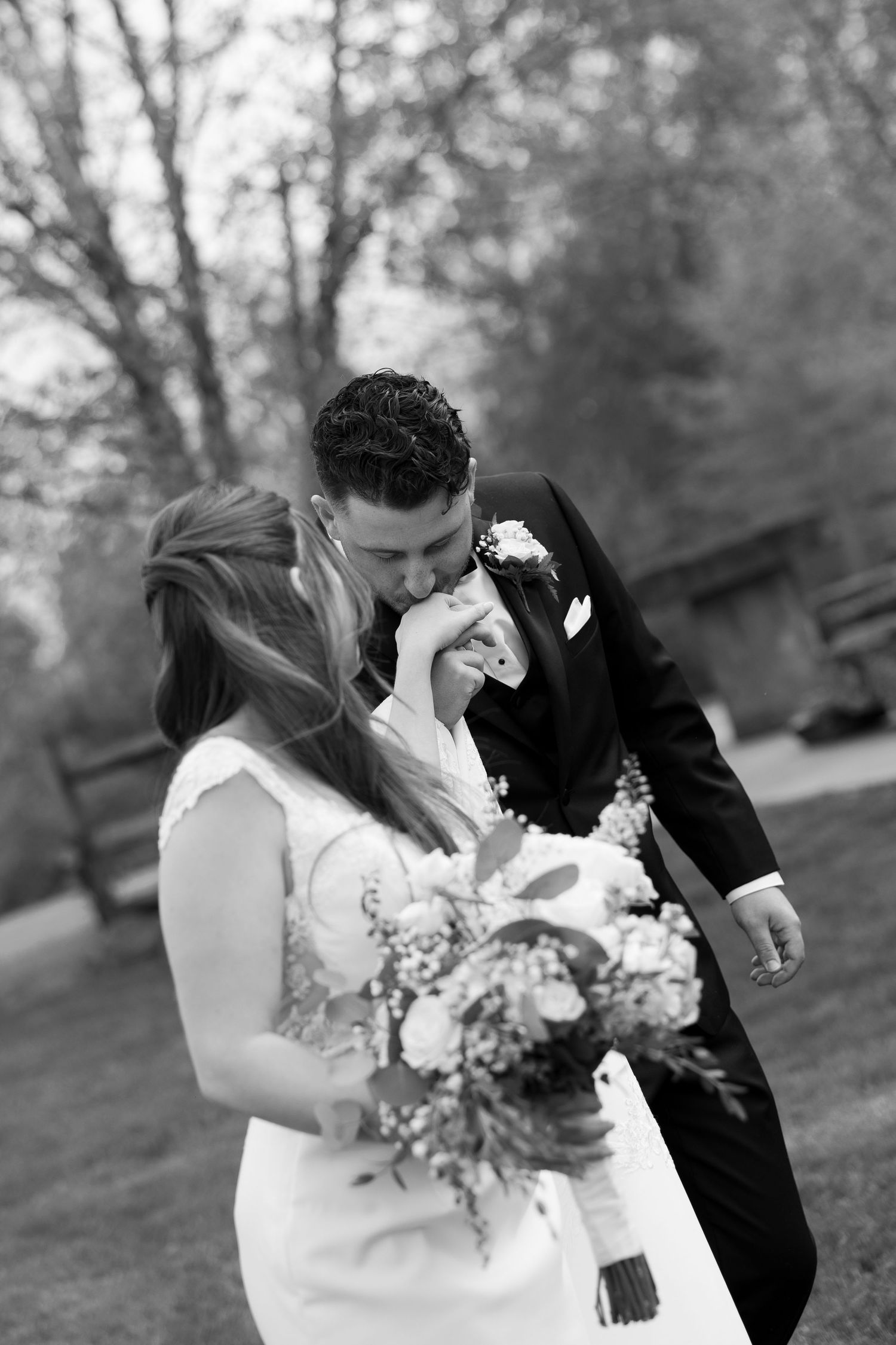 A groom tenderly kisses his emotional bride on the hand as she holds a floral bouquet in this black and white wedding portrait.
