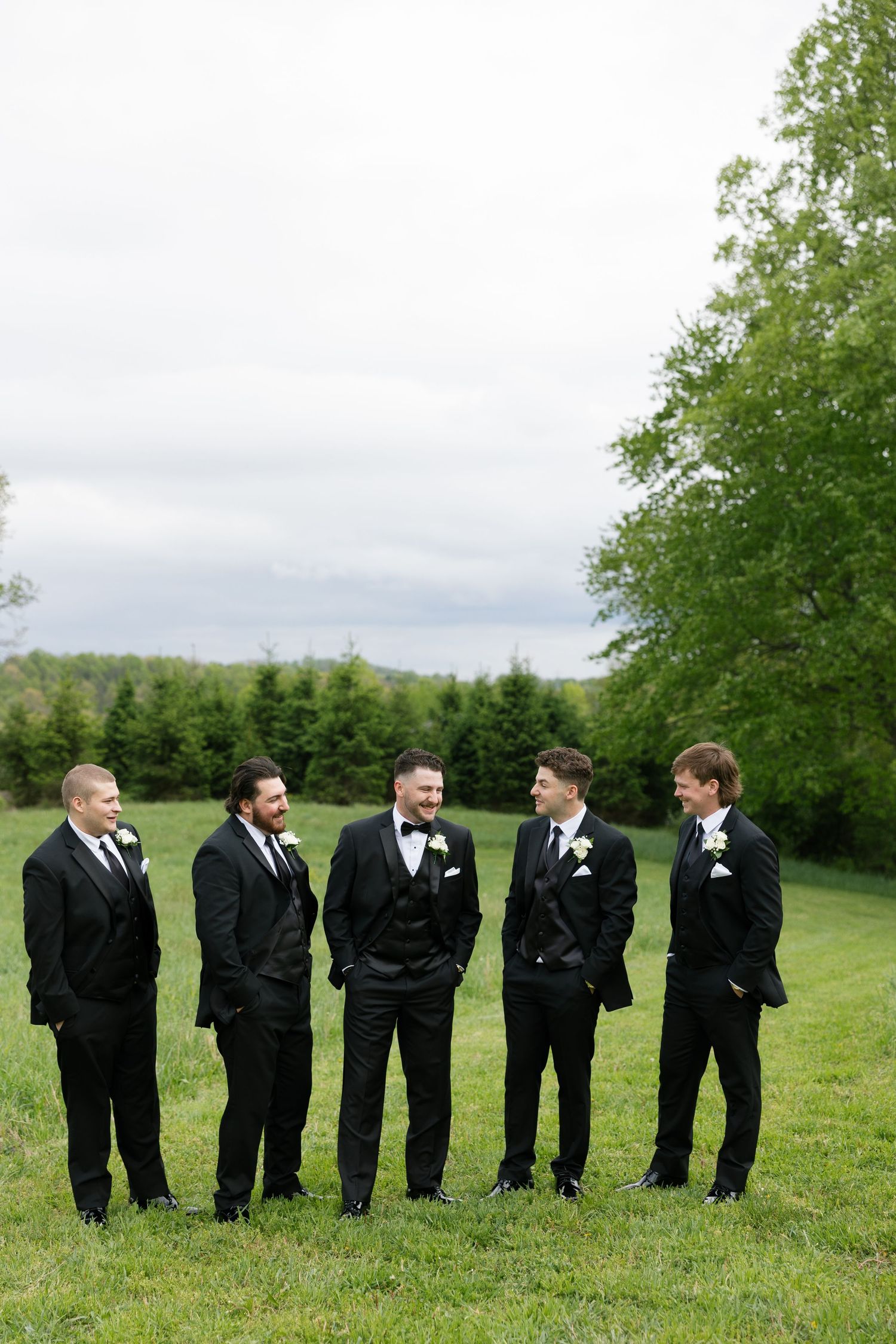 Five groomsmen in black tuxedos with white boutonnieres standing on green grass chatting outdoors.