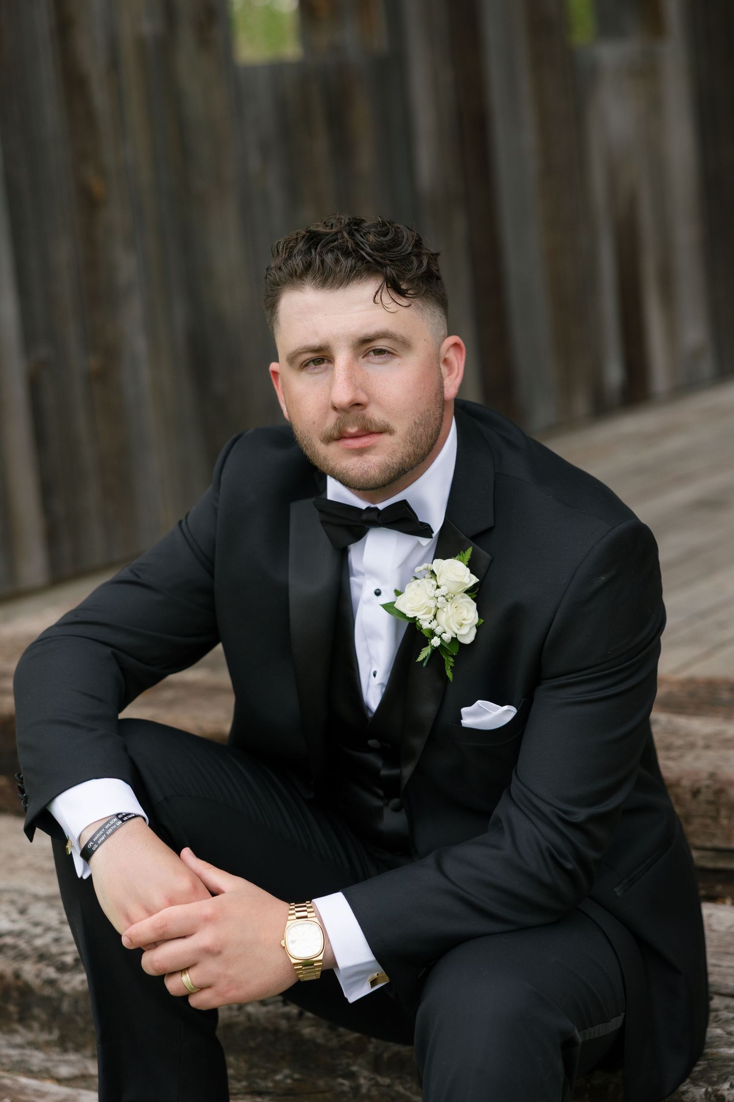 Groom in black tuxedo with white boutonniere and gold watch sitting confidently outdoors.