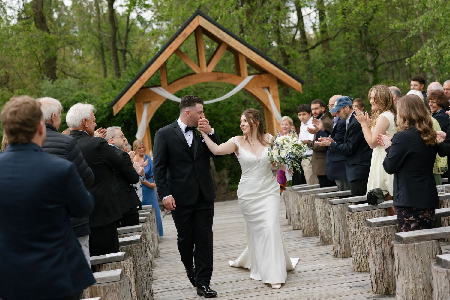 Bride and groom walking down outdoor wedding aisle under wooden arch with guests applauding.