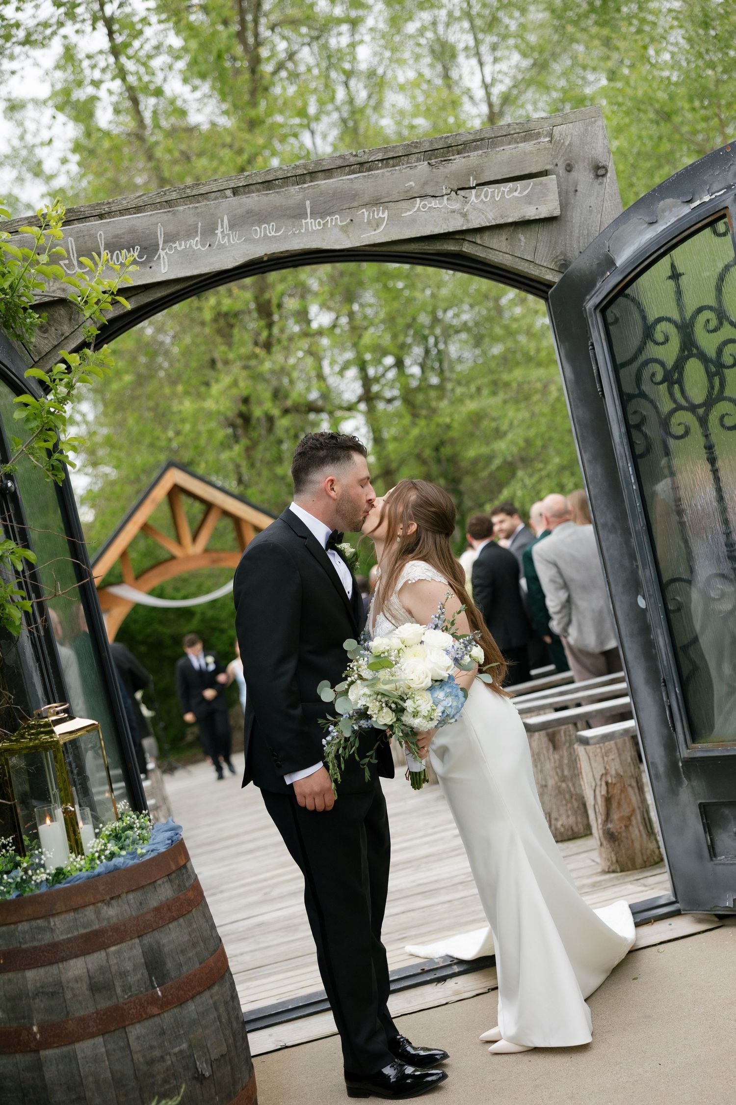 Bride and groom sharing a kiss under decorative arch with greenery at outdoor wedding venue.
