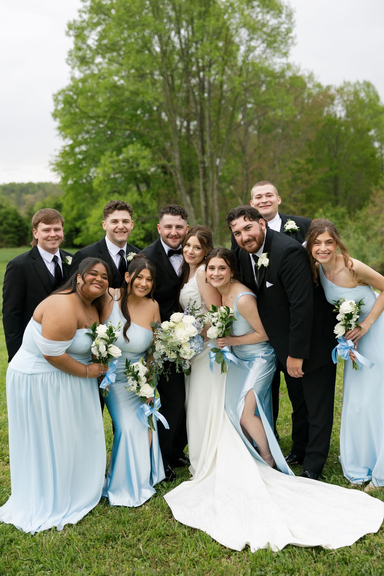 Wedding party posing outdoors with bridesmaids in light blue gowns and groomsmen in black tuxedos.