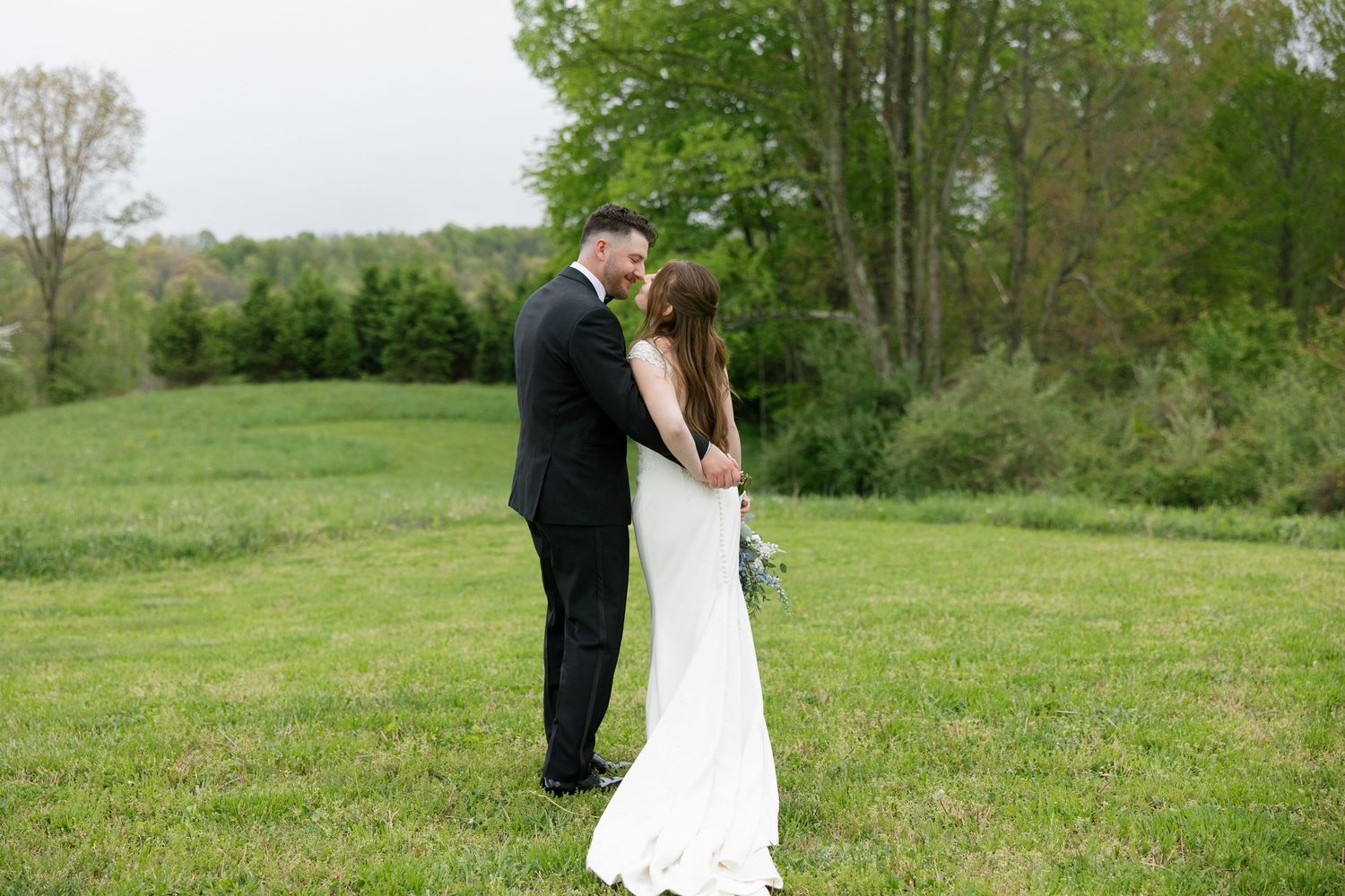 Bride and groom standing close together in an open green field surrounded by trees.
