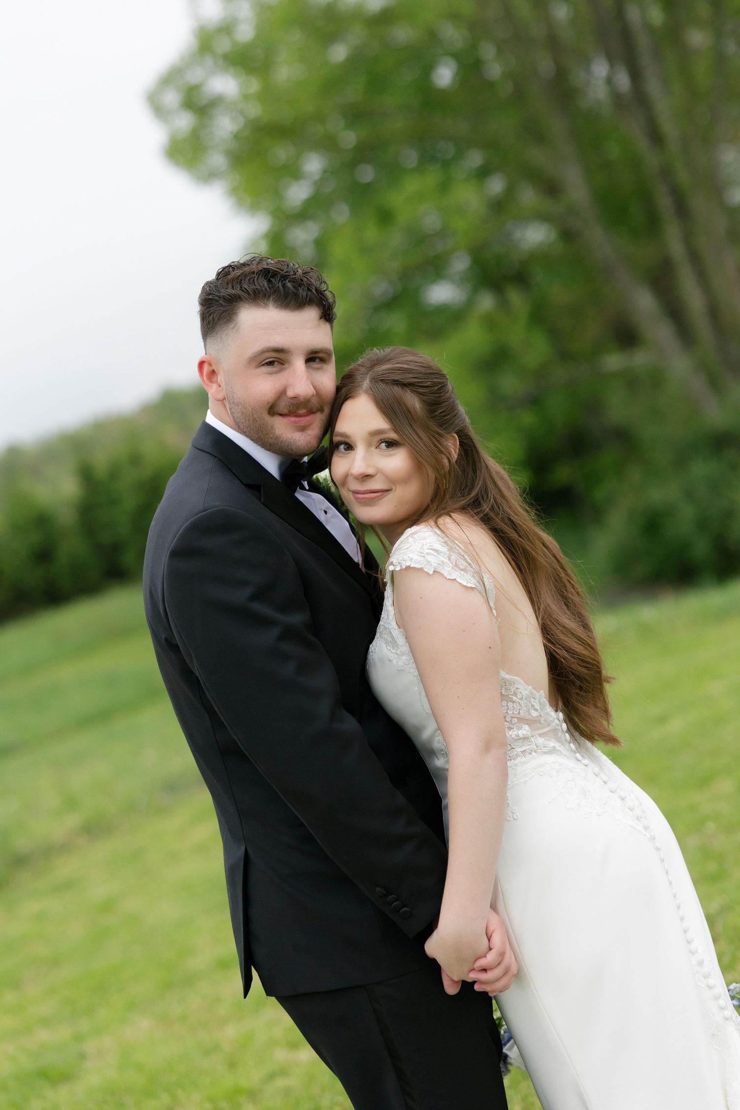 Smiling bride and groom posing together holding hands in a lush green outdoor setting.