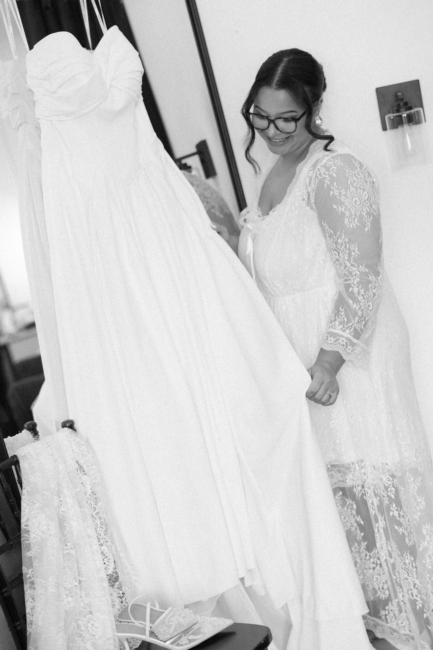 Bride in lace robe examining her white strapless wedding gown hanging in a bridal suite, black and white photo.
