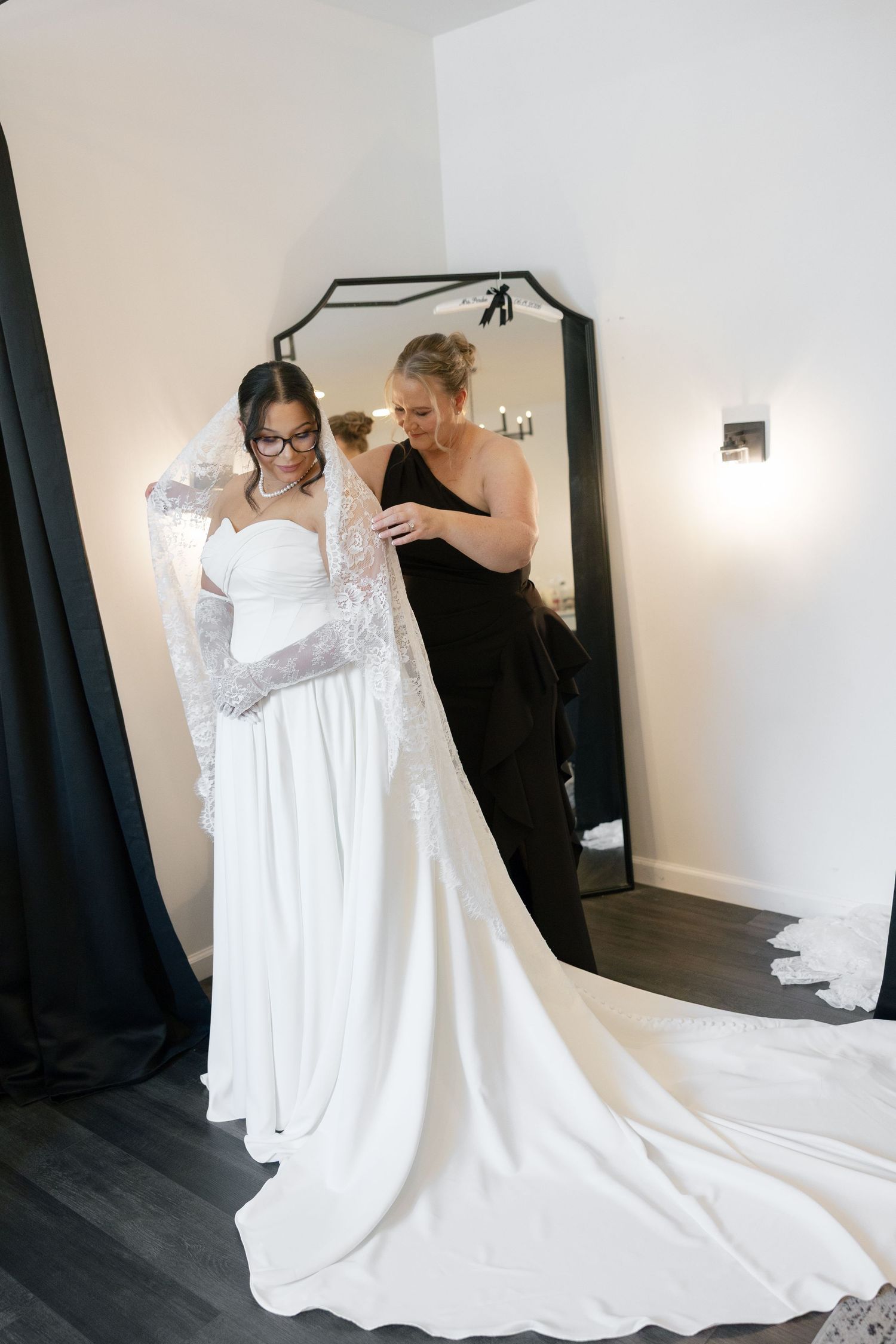 Bridesmaid in black dress helping bride into elegant white strapless wedding gown with lace veil in front of mirror.
