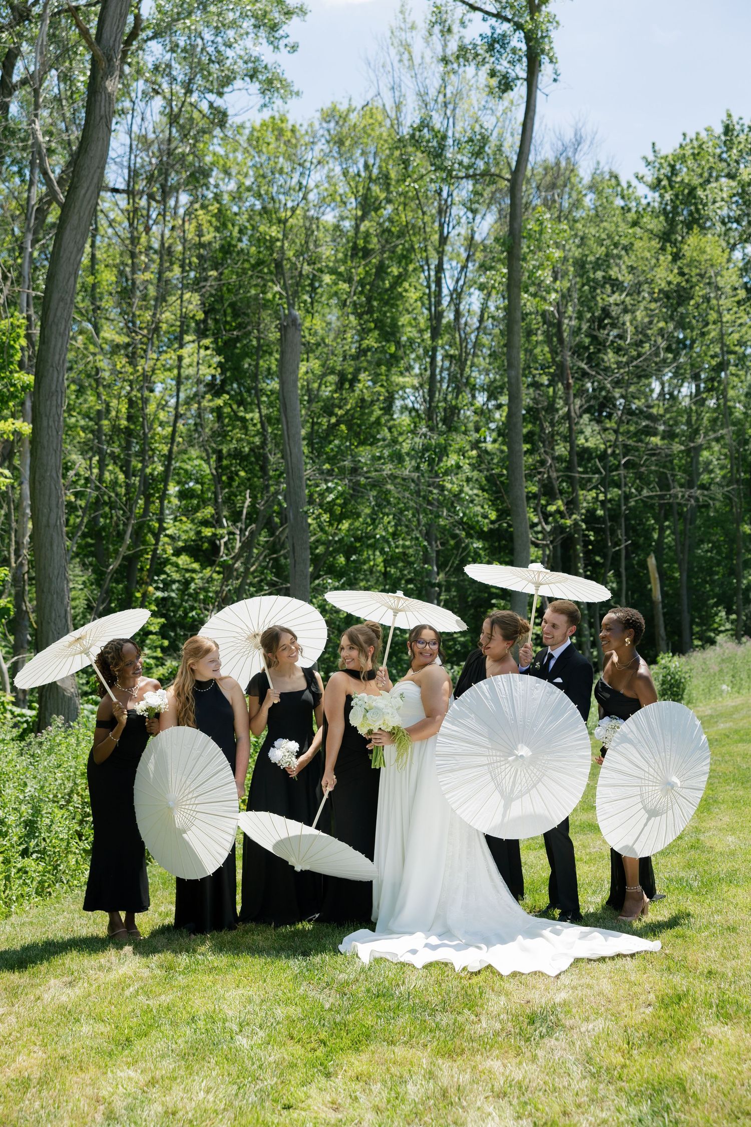 Wedding party in black attire holding white parasols with bride and groom in lush green outdoor setting.