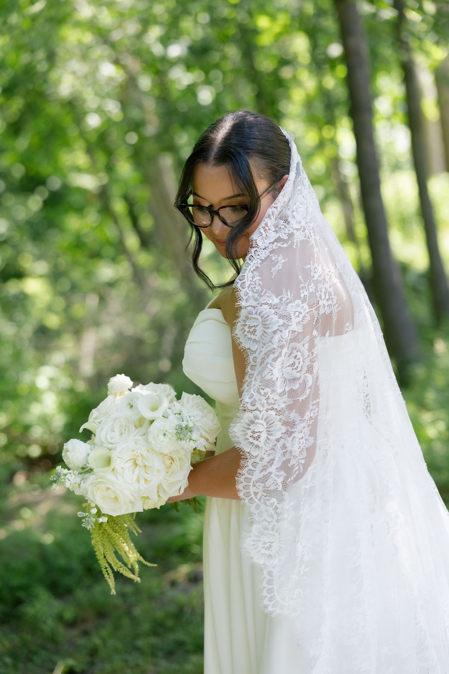 Bride wearing glasses with lace veil holding white peony bouquet in sunlit forest setting.