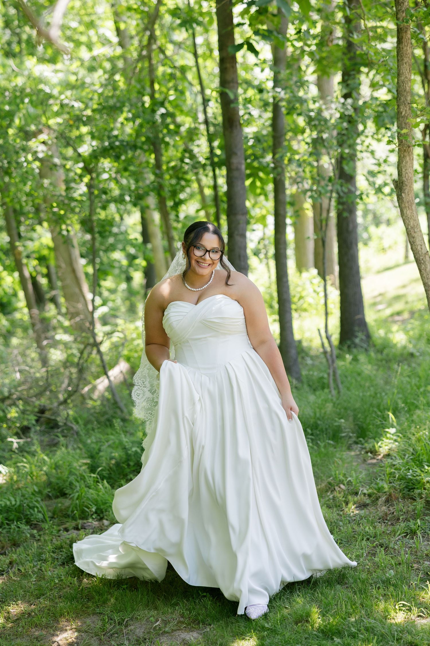 Plus-size bride in strapless white ball gown with veil and glasses smiling in green wooded setting.