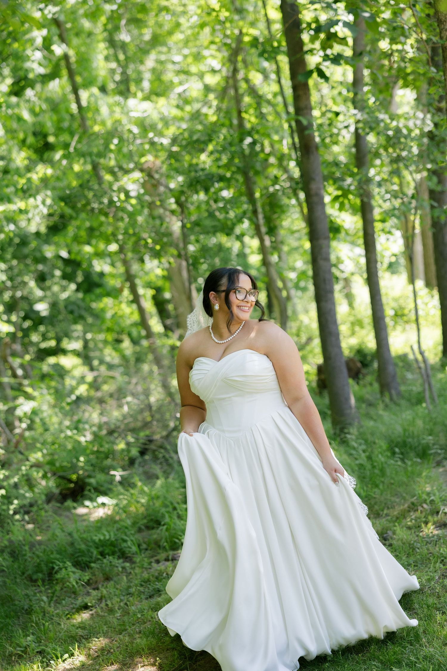 Bride in strapless white wedding gown with glasses twirling in sunlit forest, looking over shoulder.