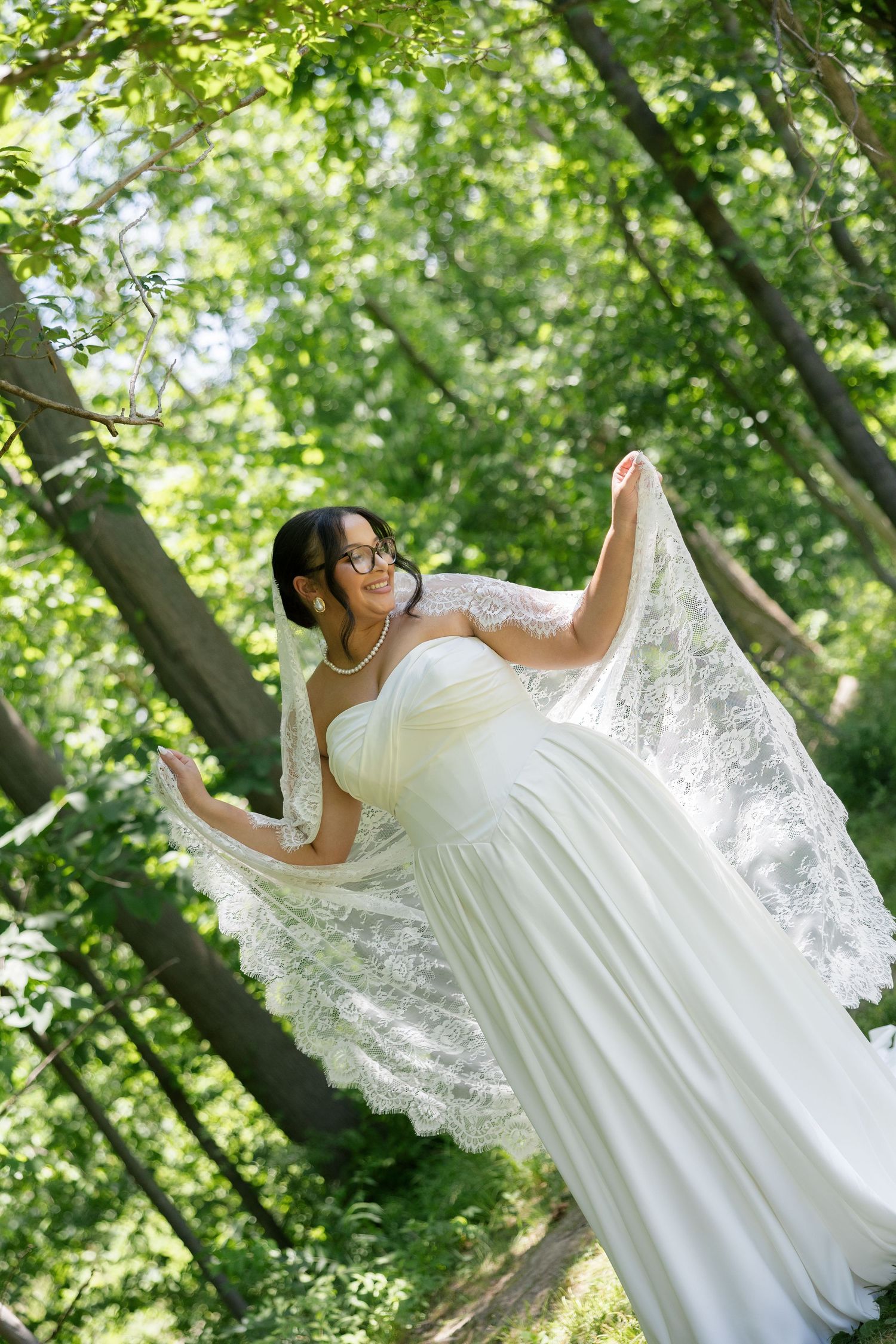Bride in strapless white gown holding lace veil open wide while posing joyfully in green forest.