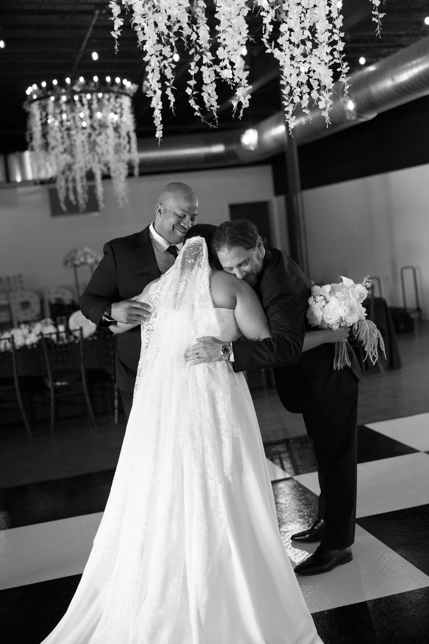 Emotional moment as two men embrace bride in lace wedding gown on checkered dance floor, black and white.