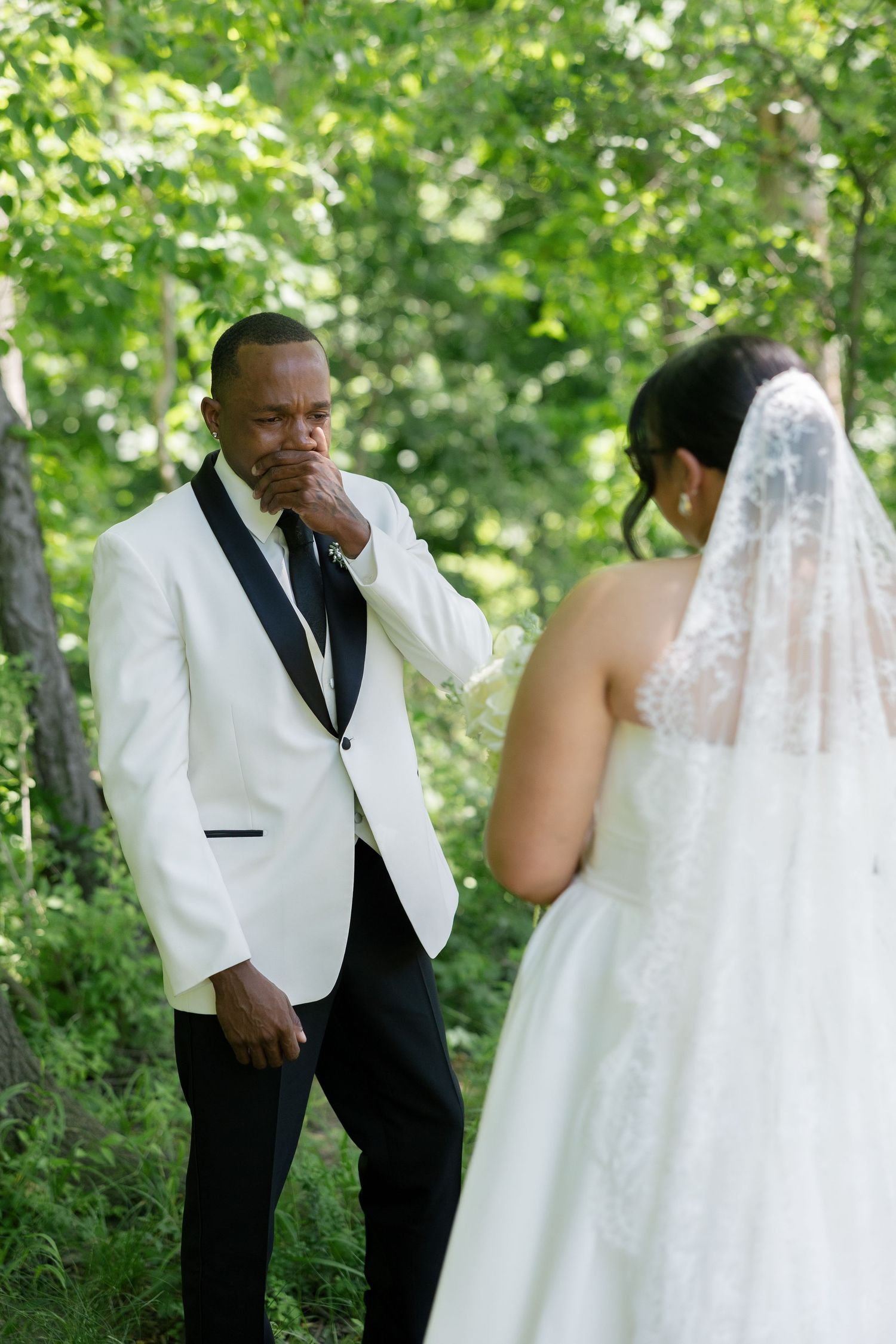 Emotional groom in white tuxedo covers mouth seeing bride in white gown for first look outdoors.