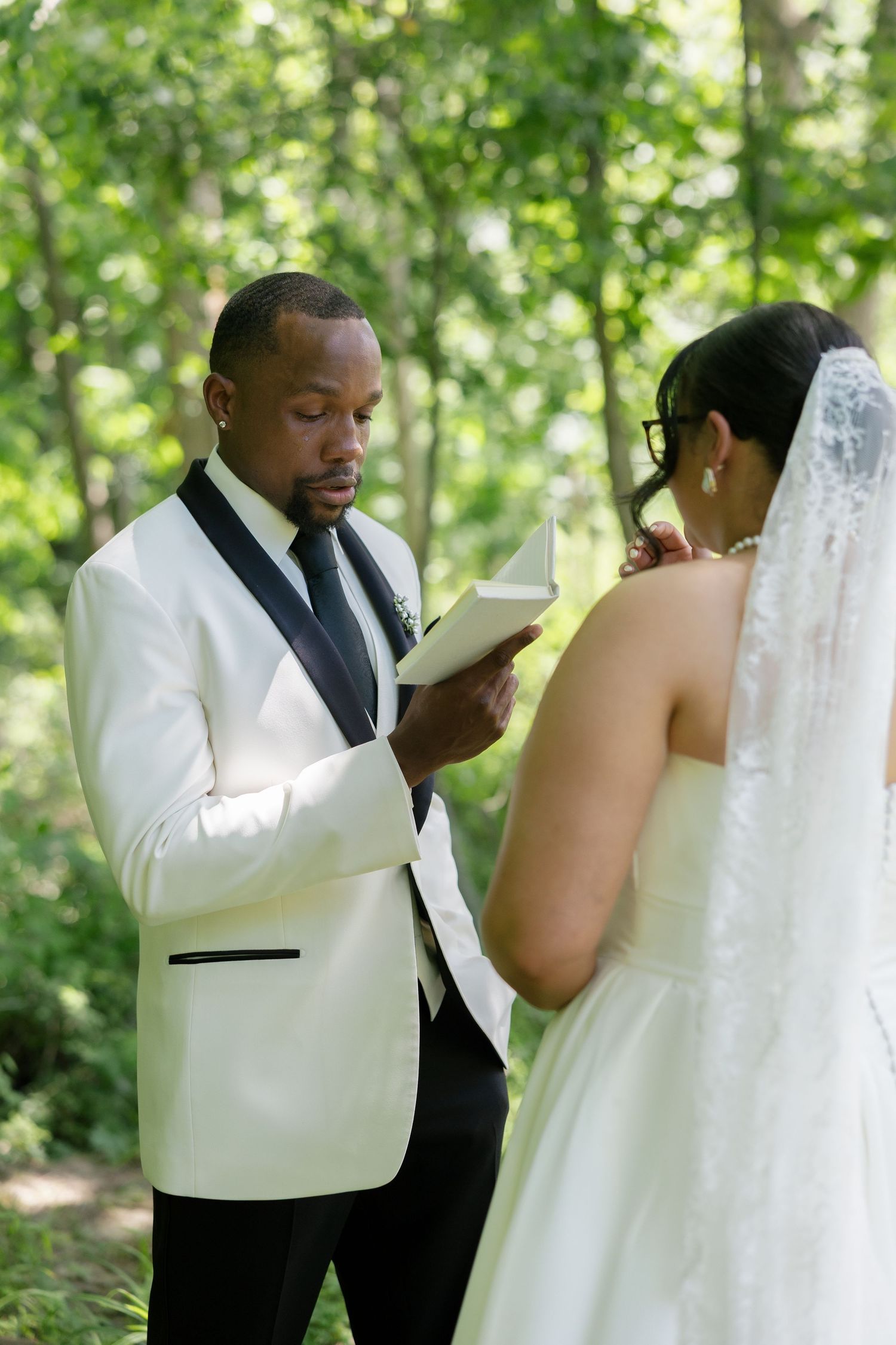 Groom in white tuxedo reads handwritten vows to bride in lace veil during outdoor forest ceremony.
