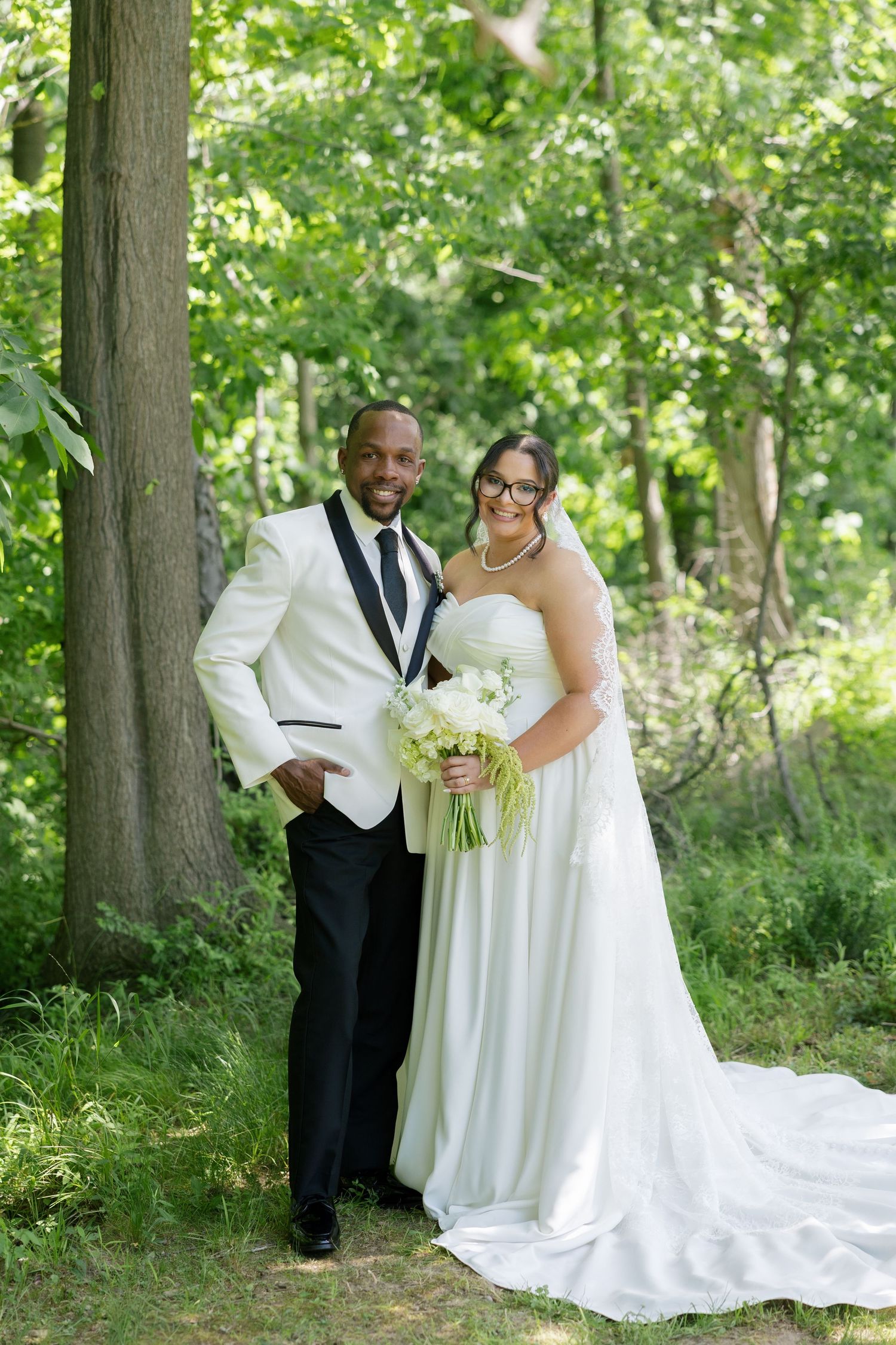 Bride and groom pose together in lush green forest, she holds white bouquet in strapless gown.
