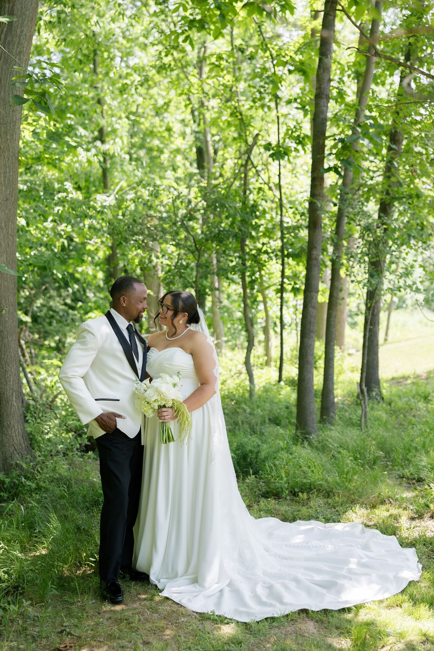 Bride and groom share romantic forehead touch moment surrounded by tall trees in sunlit forest.