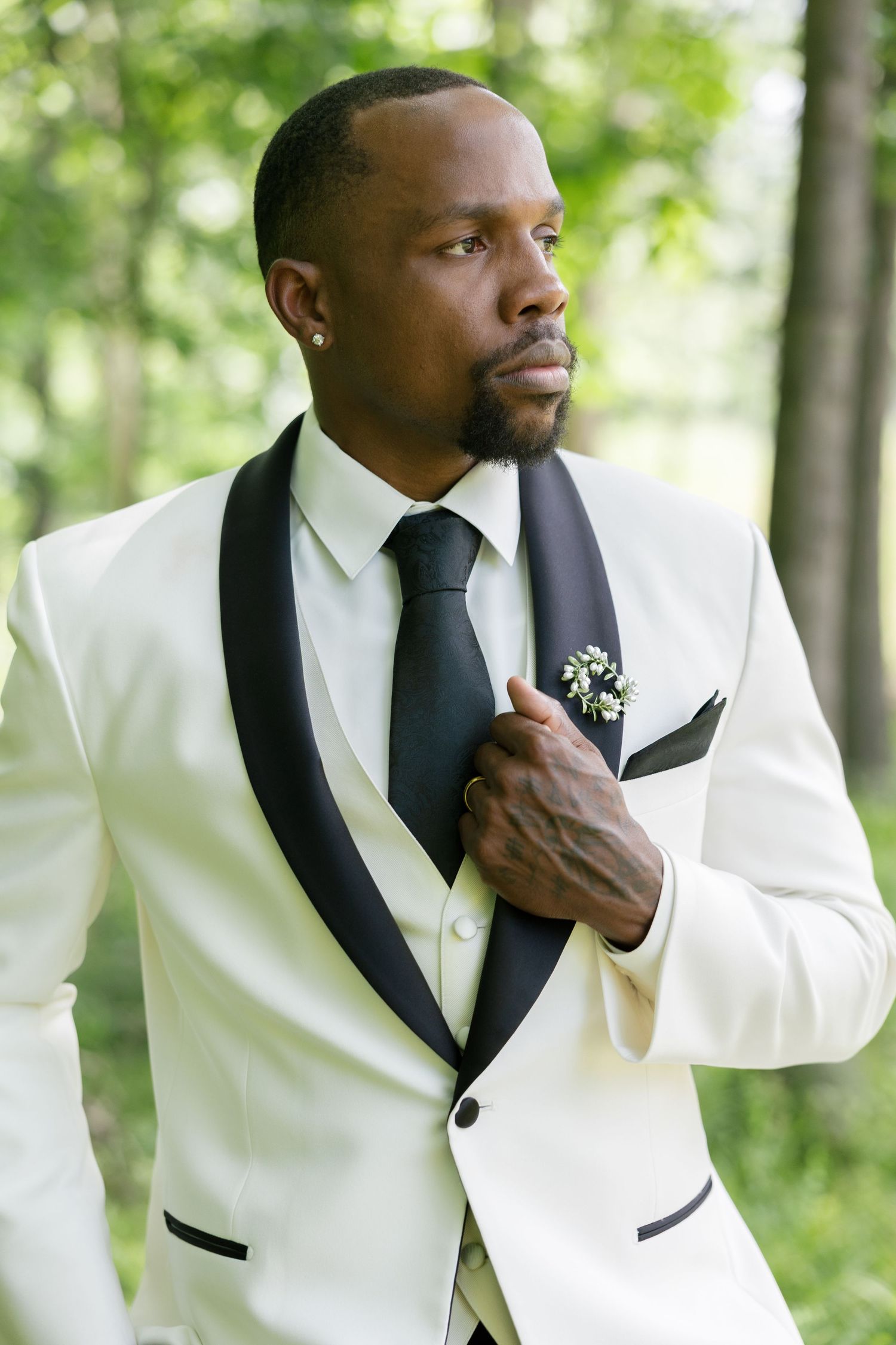 Handsome groom in white tuxedo with black lapels and boutonniere posing outdoors in wooded setting.