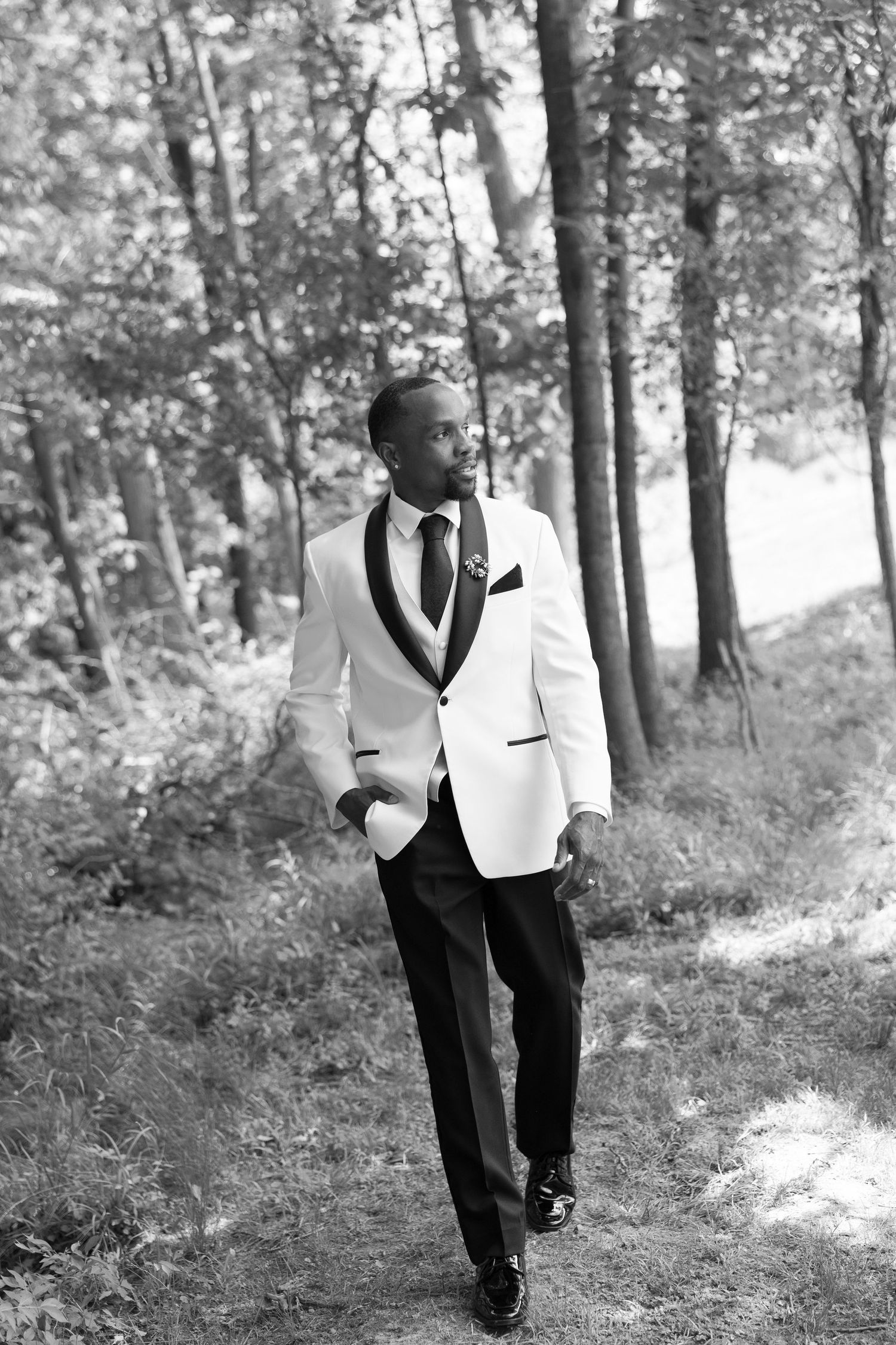 A dapper groom in a white tuxedo jacket and black trousers walks through a wooded forest path in this black and white portrait.