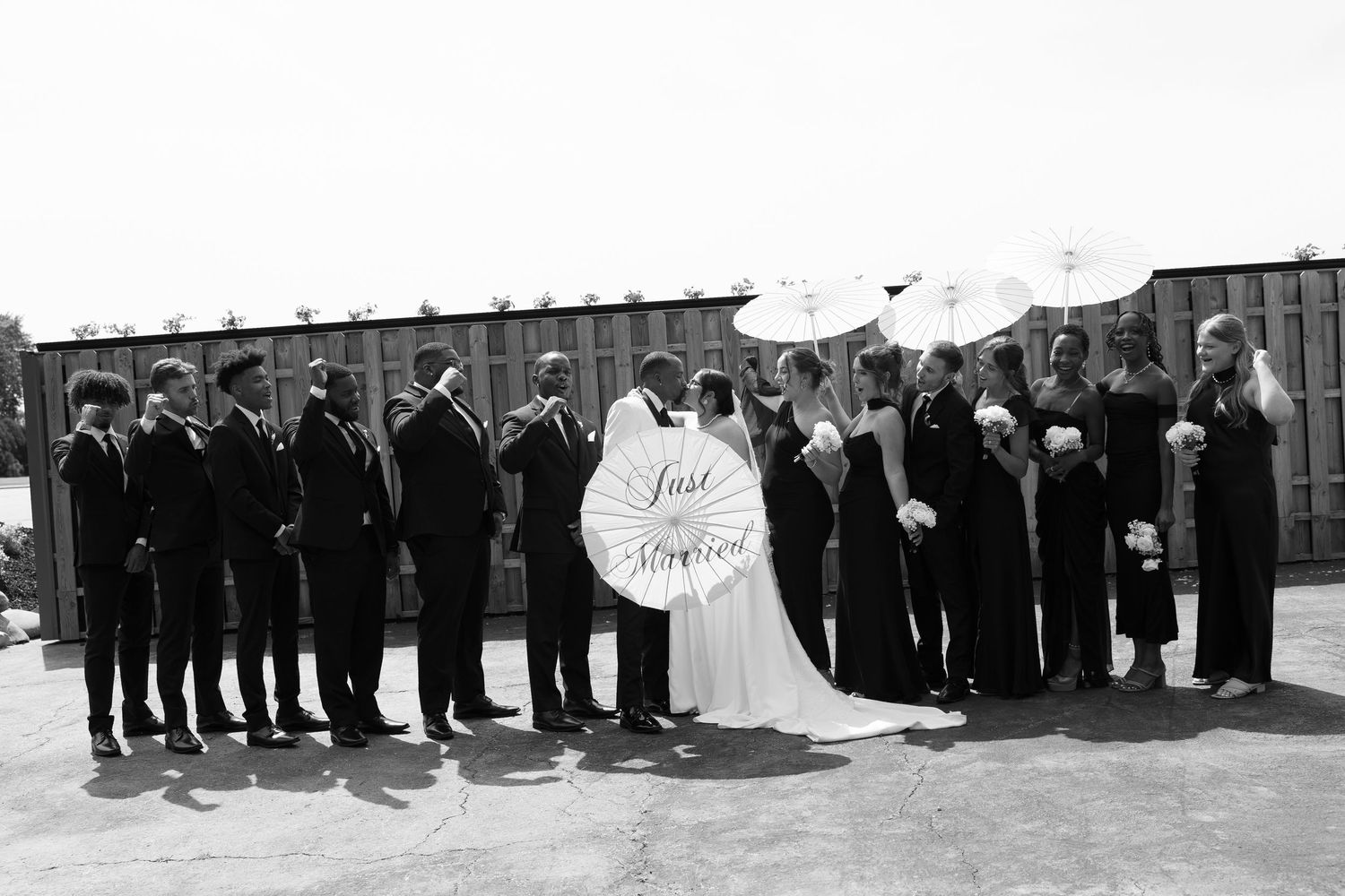 Wedding party posing in black and white with parasols and Just Married sign outside industrial venue.