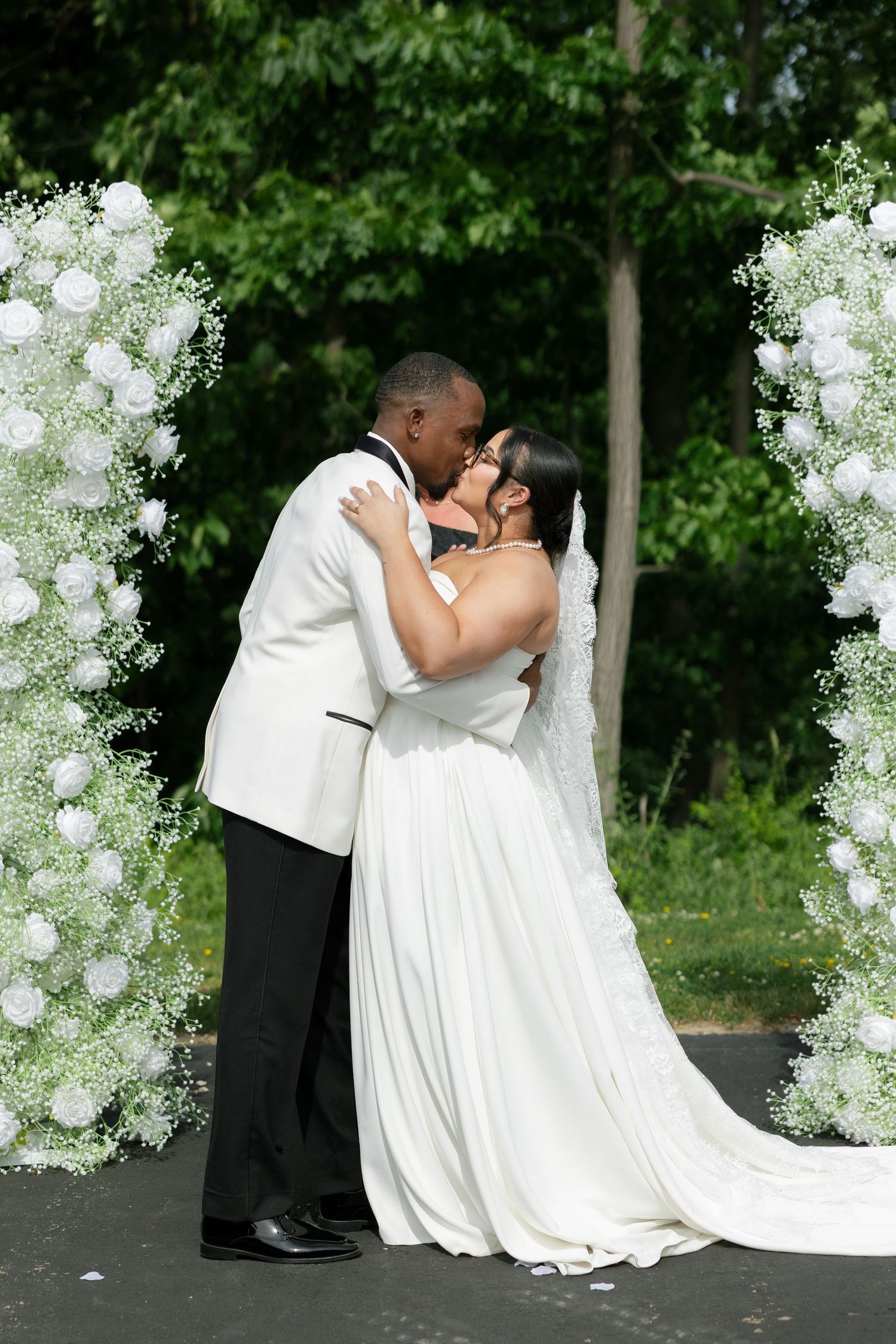 A bride and groom share their first kiss under a floral arch adorned with white roses and baby's breath at an outdoor wedding ceremony.