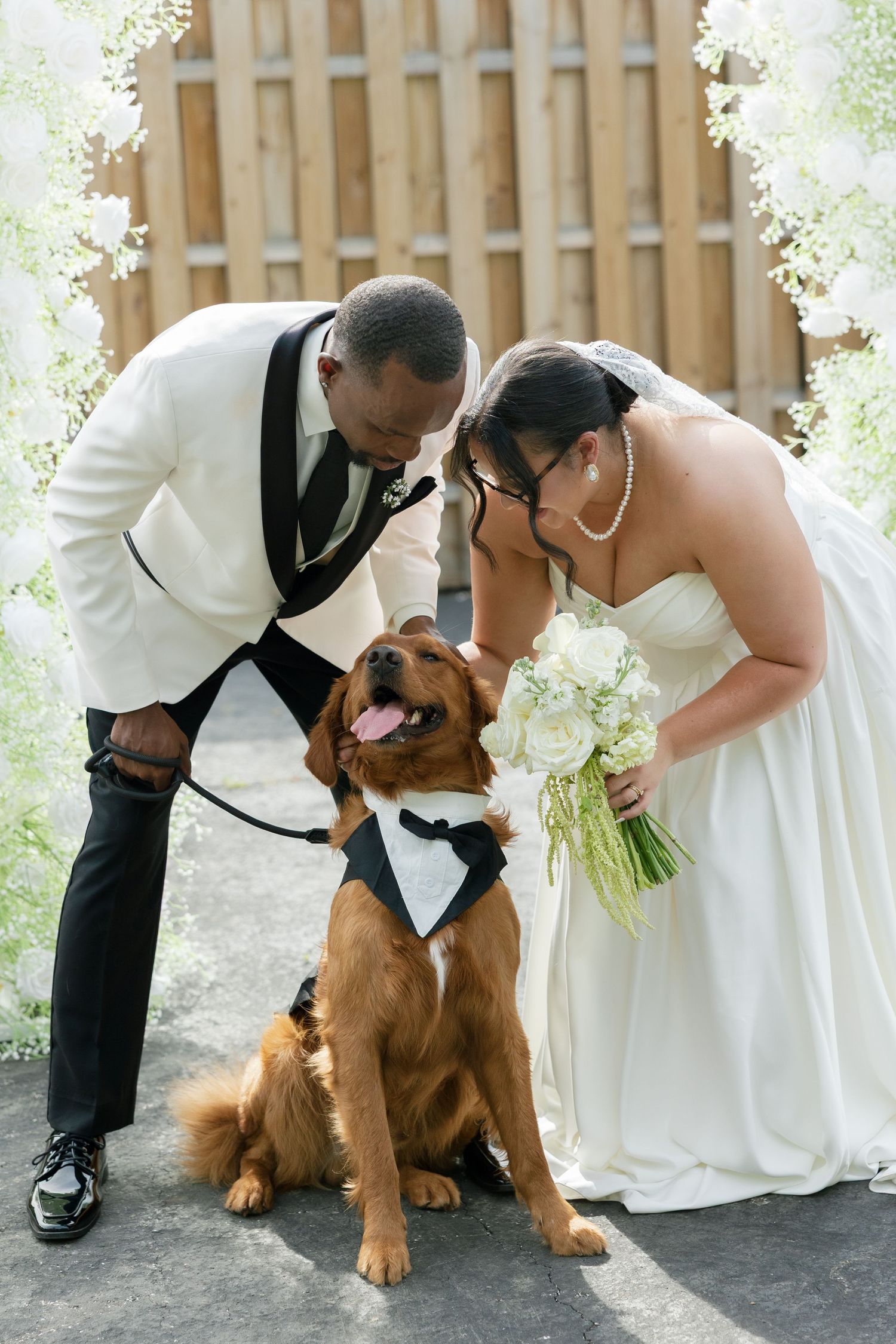 A newlywed couple in wedding attire bends down to pet their golden retriever wearing a tuxedo bandana at an outdoor ceremony.