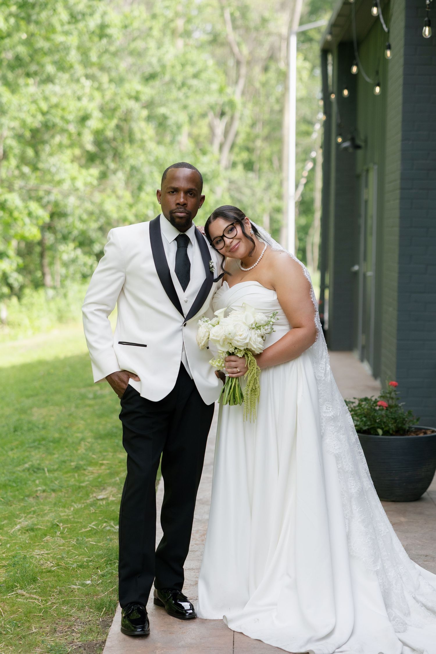 Elegant bride and groom pose outside green building, she leans on his shoulder holding white bouquet.