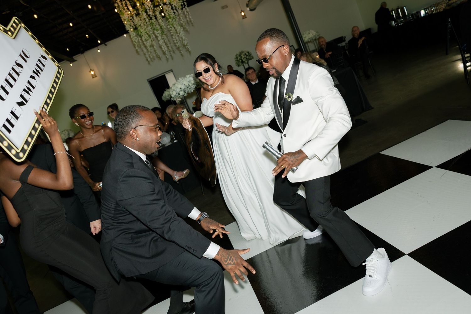 A bride and groom in a white tuxedo jacket celebrate with guests on a black and white checkered dance floor at their wedding reception.