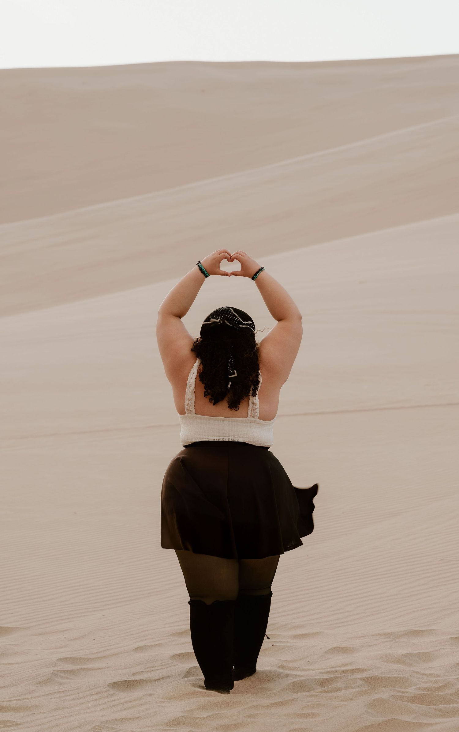 Woman in white top and black skirt forms heart shape with hands above head while standing on sandy desert dunes.