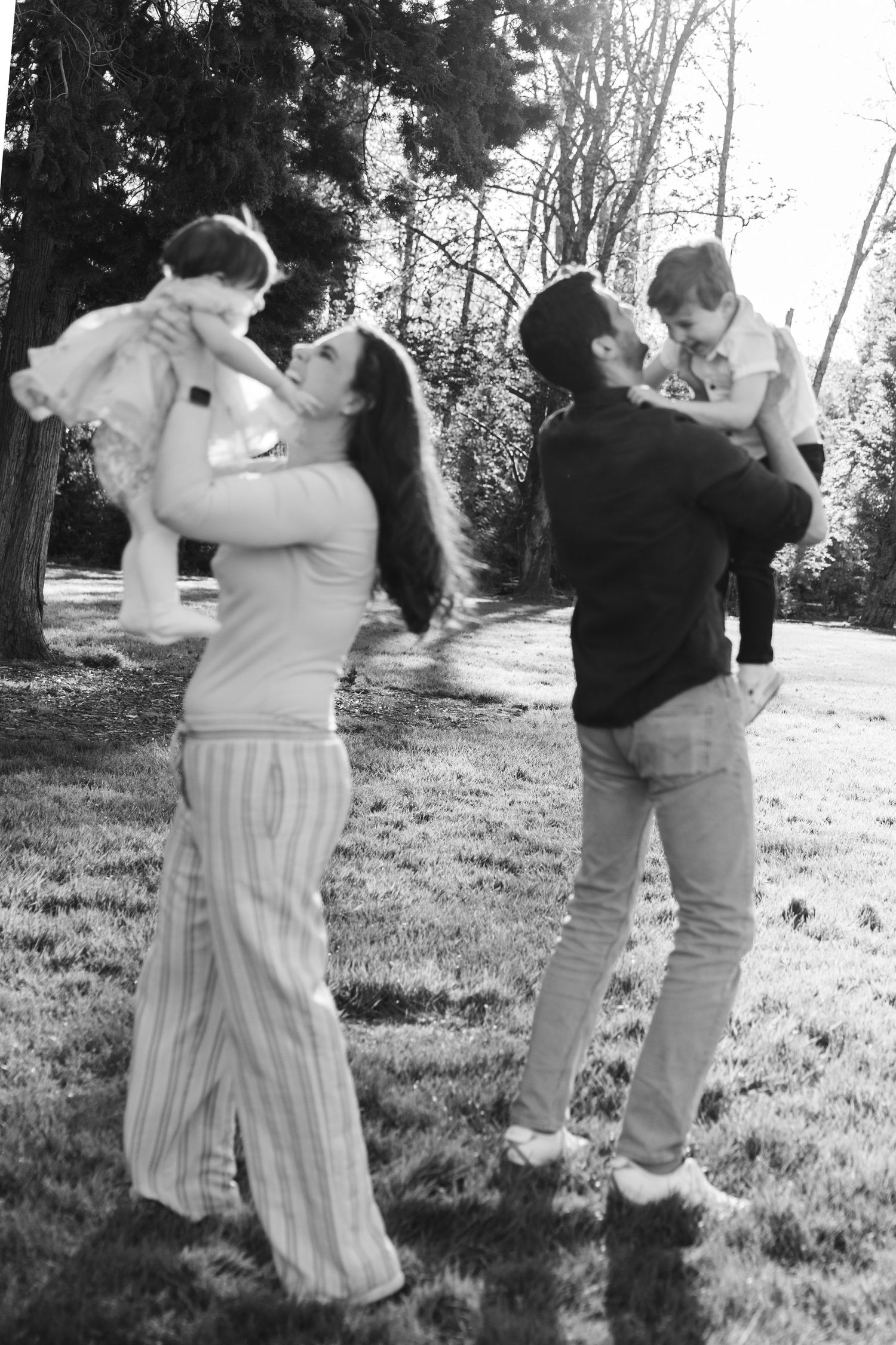 Black and white photo of two parents lifting their babies in a sunny park, sharing a joyful family moment outdoors.