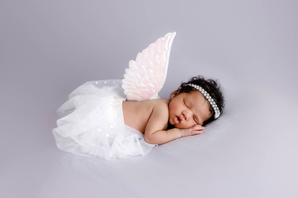 Newborn baby girl sleeping peacefully in white tutu dress with pink angel wings and pearl headband.