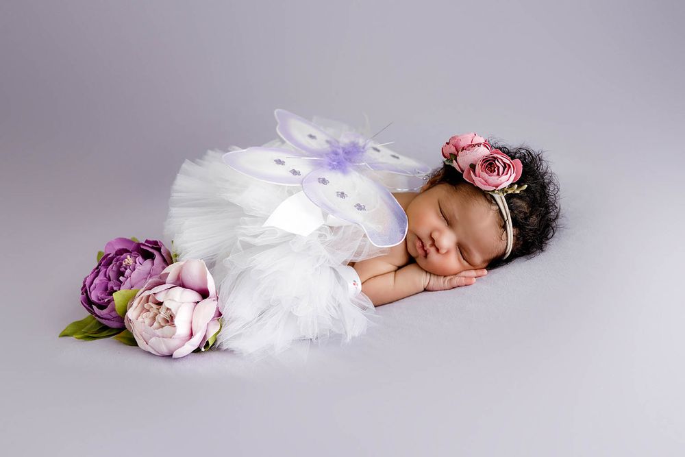 Sleeping newborn baby dressed in white fairy costume with butterfly wings and floral headband surrounded by purple and pink flowers.