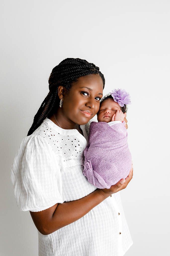 A smiling mother with braids holds her swaddled newborn in a purple wrap and floral headband against a white background.